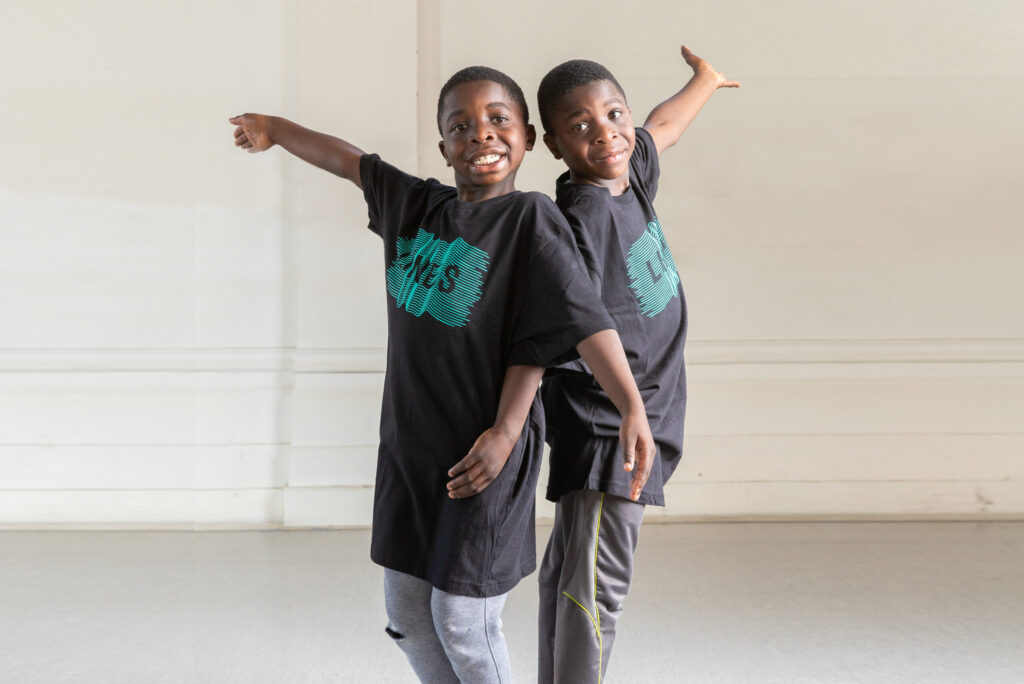 Two kids at LINES Dance Center standing back to back in LINES shirts, holding their arms out to the sides and smiling