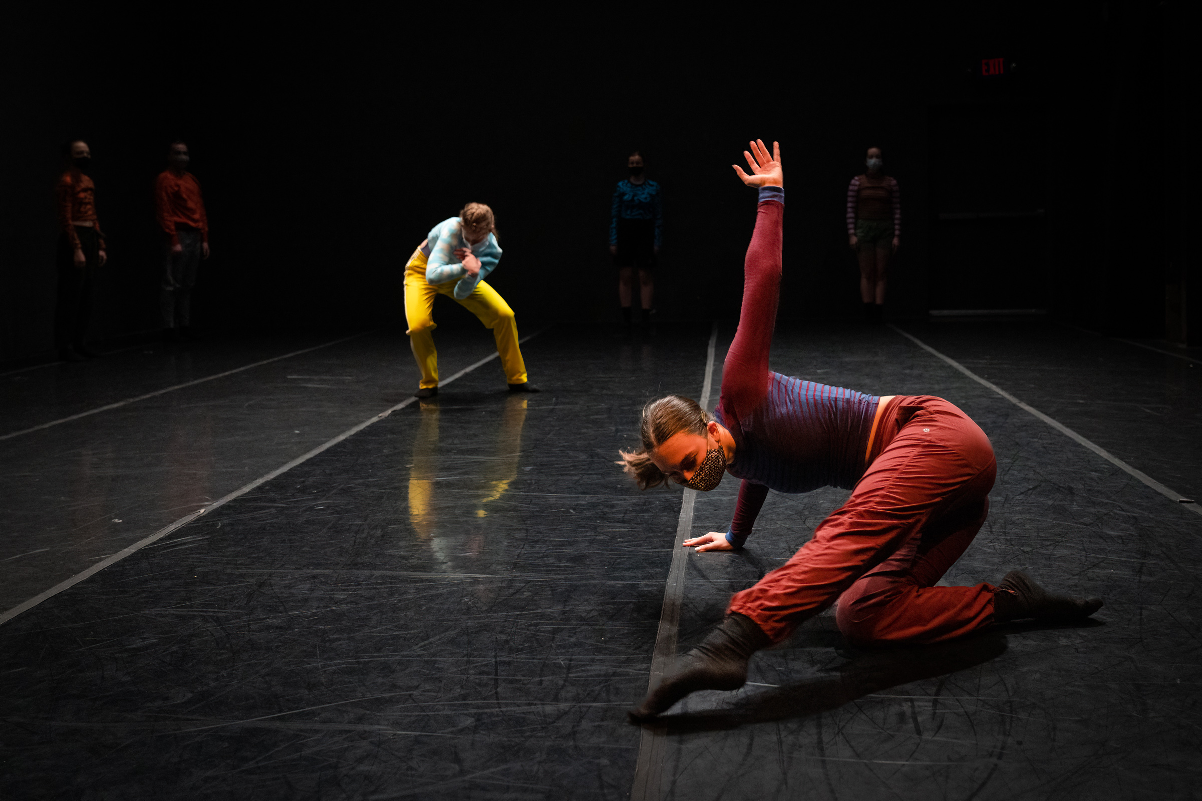 LINES Ballet | Training Program artists performing a work by Brett Conway on stage, one student is doing work in red pants and blue top with red sleeves while the other stands with spine curled, arms in, and legs bent while wearing yellow pants and light blue shirt, other dancers can be seen further back standing on stage, all dancers are wearing masks