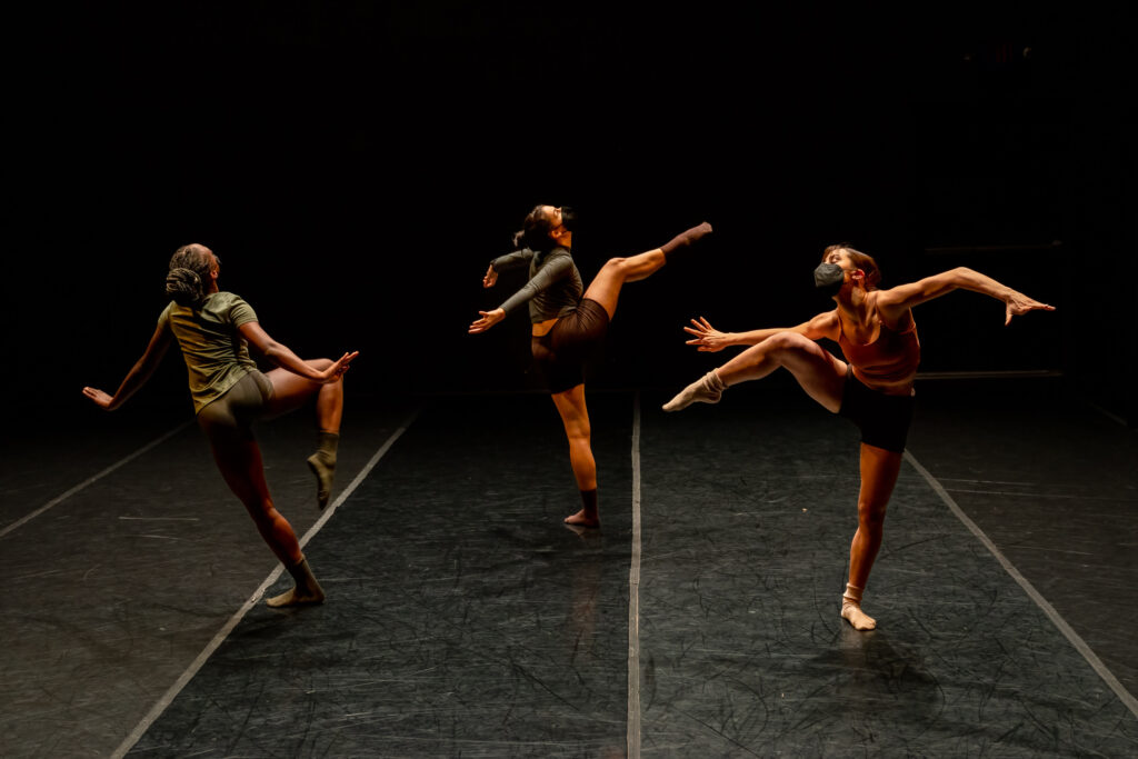 Three LINES Ballet | Training Program artists performing a work by Erik Wagner on stage, two are facing the back, one is facing the front, all dancers are wearing masks and have their legs in side attitude, they are falling away from the lifted leg while arms push and stretch