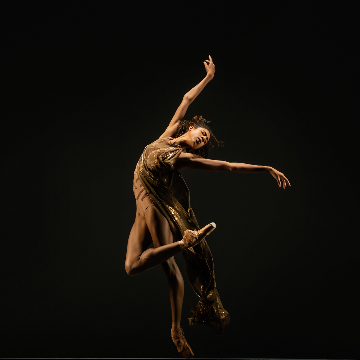 LINES Ballet company artist Adji Cissoko en pointe leaning over the edge of one pointe shoe with the other leg bent at the knee and tucked across the front of the standing leg. Arms are sweeping, one up above the body, the other out to the side.