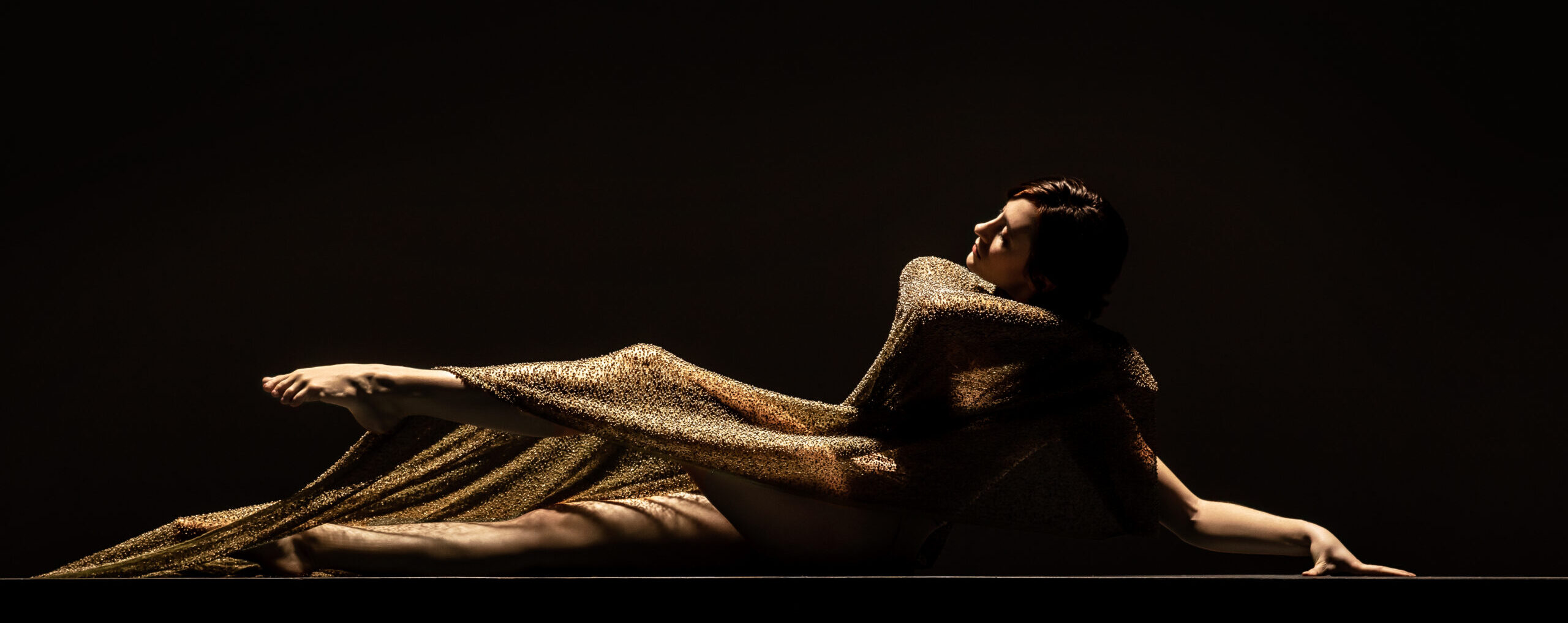 LINES Ballet company artist Maya Harr sitting on the floor draped in a glittering gold sheer sheet of fabric, legs outstretched in front, one lifted slighting off the ground with pointed foot, one arm reaching behind and touching the floor for support and other arm wrapping across the opposite shoulder