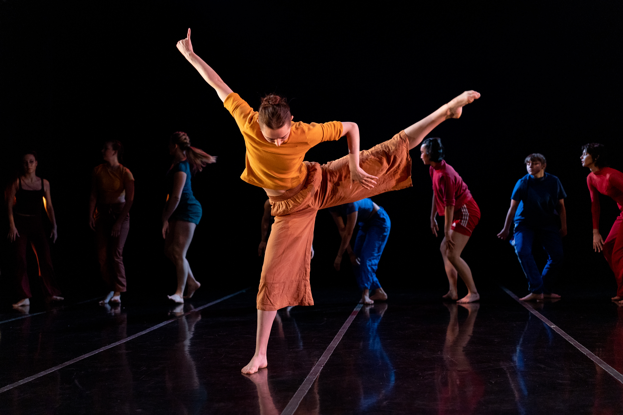 LINES Ballet Training Program dancers performing on stage in a showcase; a dancer in orange, who is holding their leg up to the side, is the focal point.