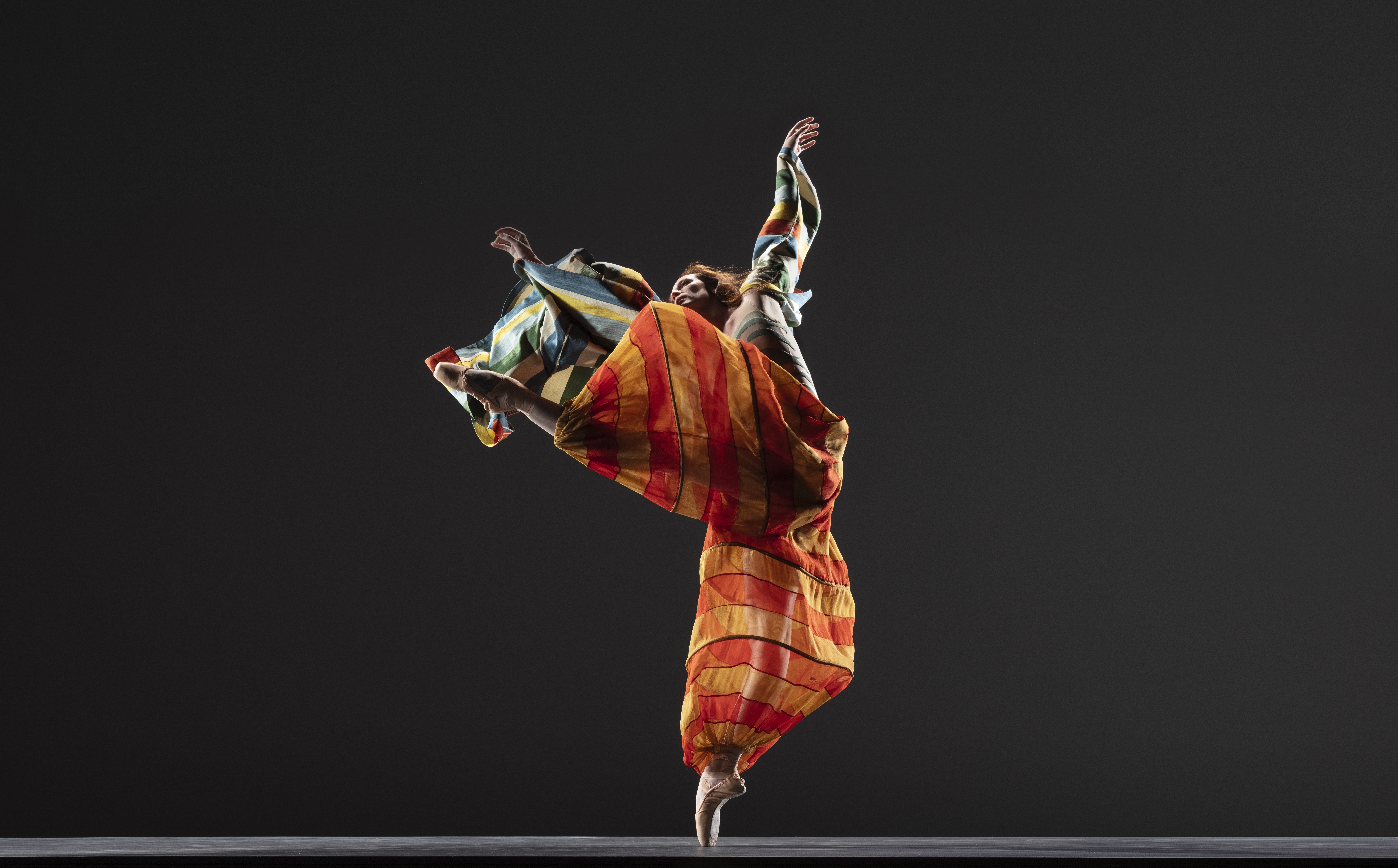 LINES Ballet company dancer Madeline DeVries dancing en pointe wearing red and yellow striped flowy pants and a red and blue striped flowy shirt; one leg is extended out, as the arms extend