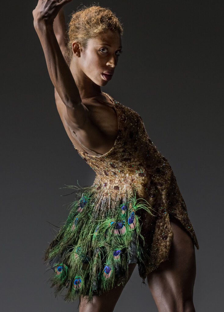 LINES Ballet company dancer Adji Cissoko looking over her shoulder towards the camera, wearing a dress made of peacock feathers with a gold bodice; Adji has her arms raised over her head