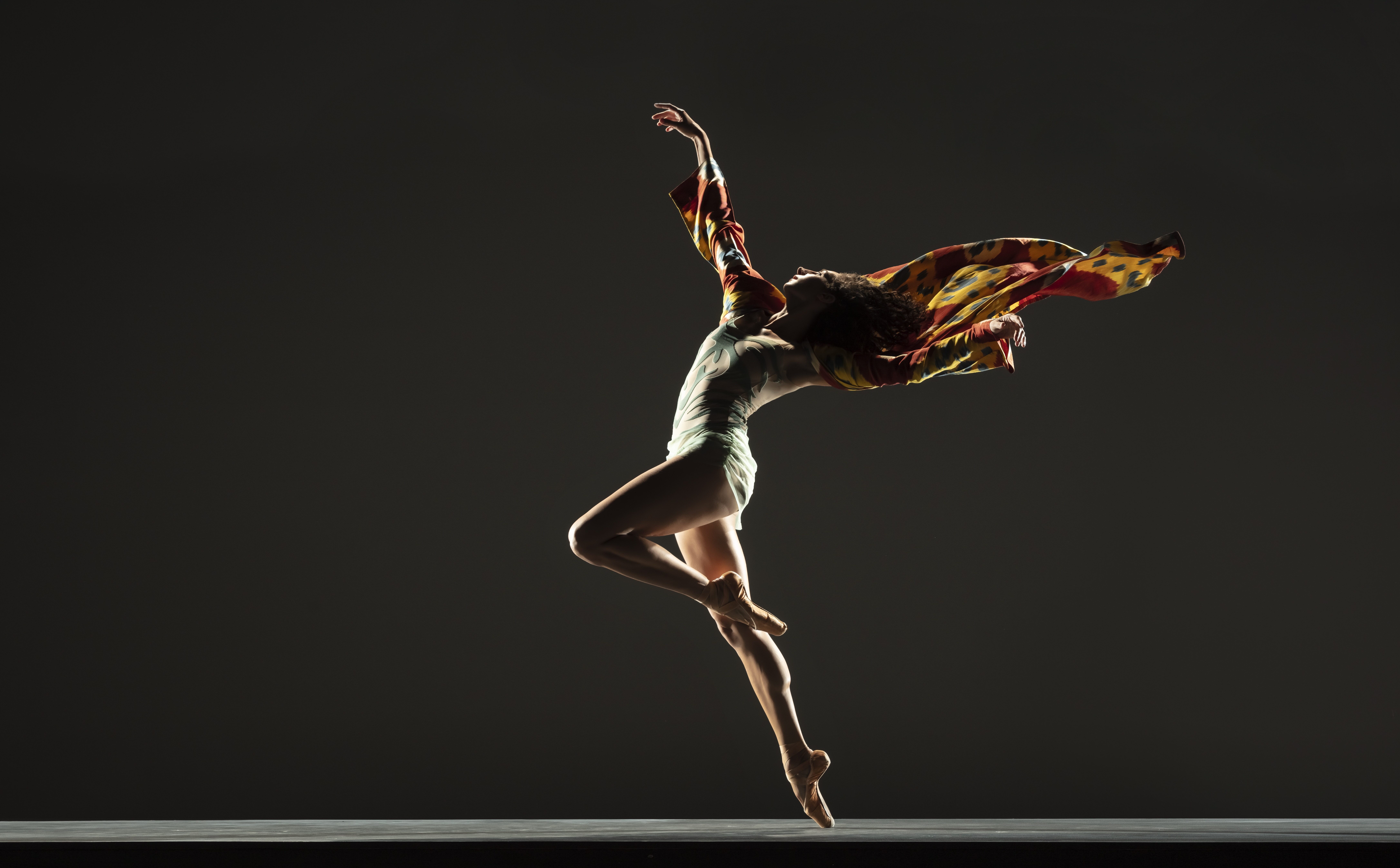 LINES Ballet company dancer Ilaria Guerra in passé en pointe, back arched and arms extended as a maroon and yellow cape wafts behind her shoulders
