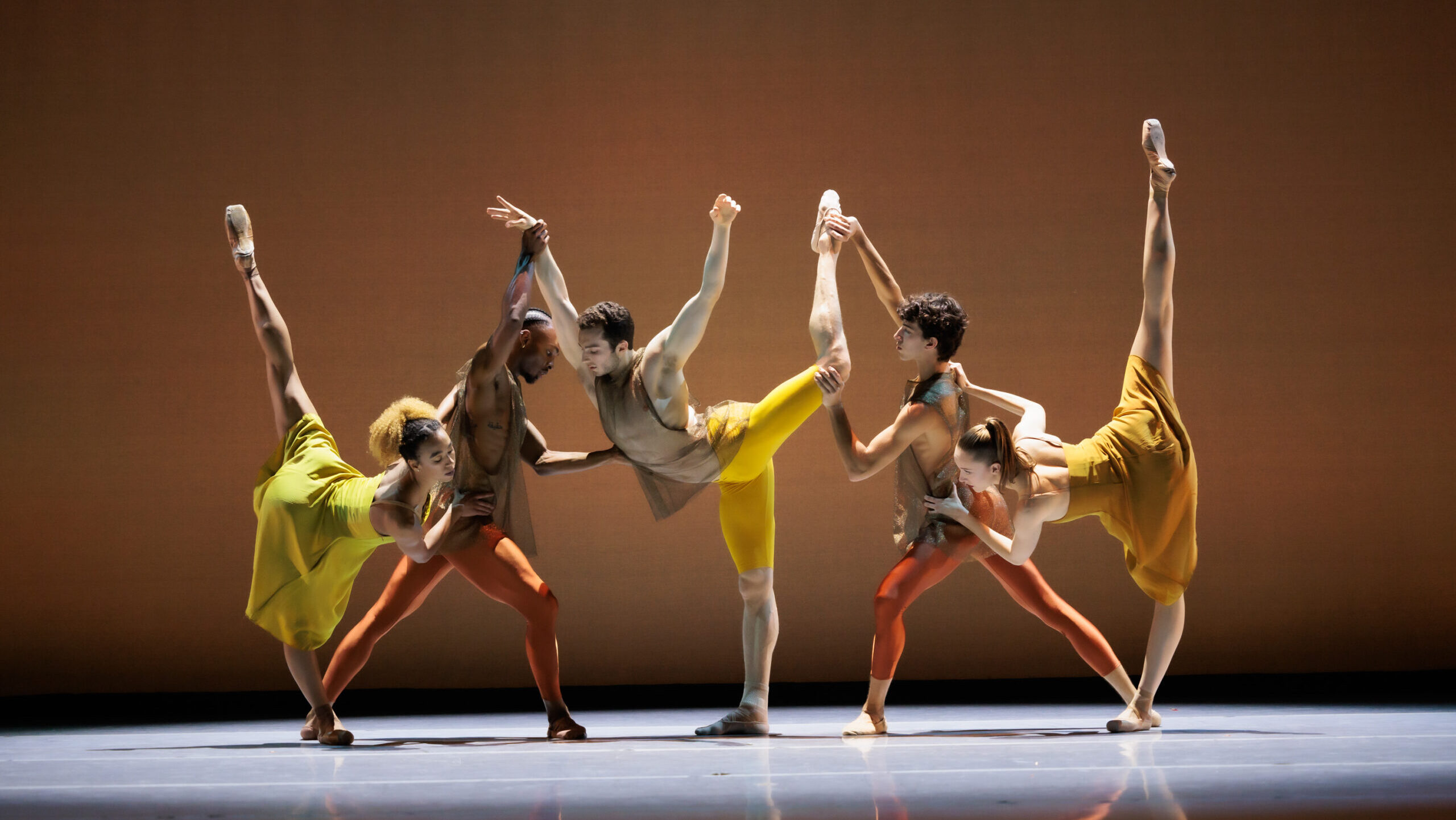 LINES Ballet Dancers Marusya Madubuko, Joshua Francique, Theo Duff-Grant, Maël Amatoul, and Tatum Quiñónez connect in a line onstage, various limbs reaching up and out, all connected to one another. Costumes are bright yellows, oranges, and browns.