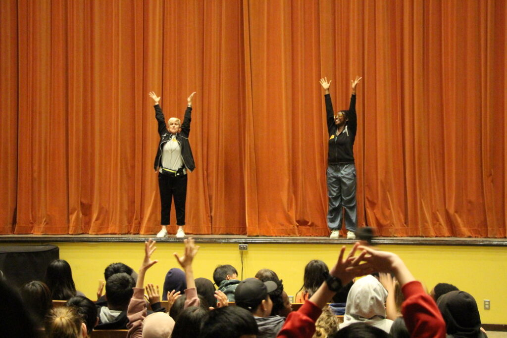 Mary Carbonara and another educator standing on stage leading an auditorium of students at Aptos Middle School in movement before the LINES Ballet company dancers perform in an on-site school performance