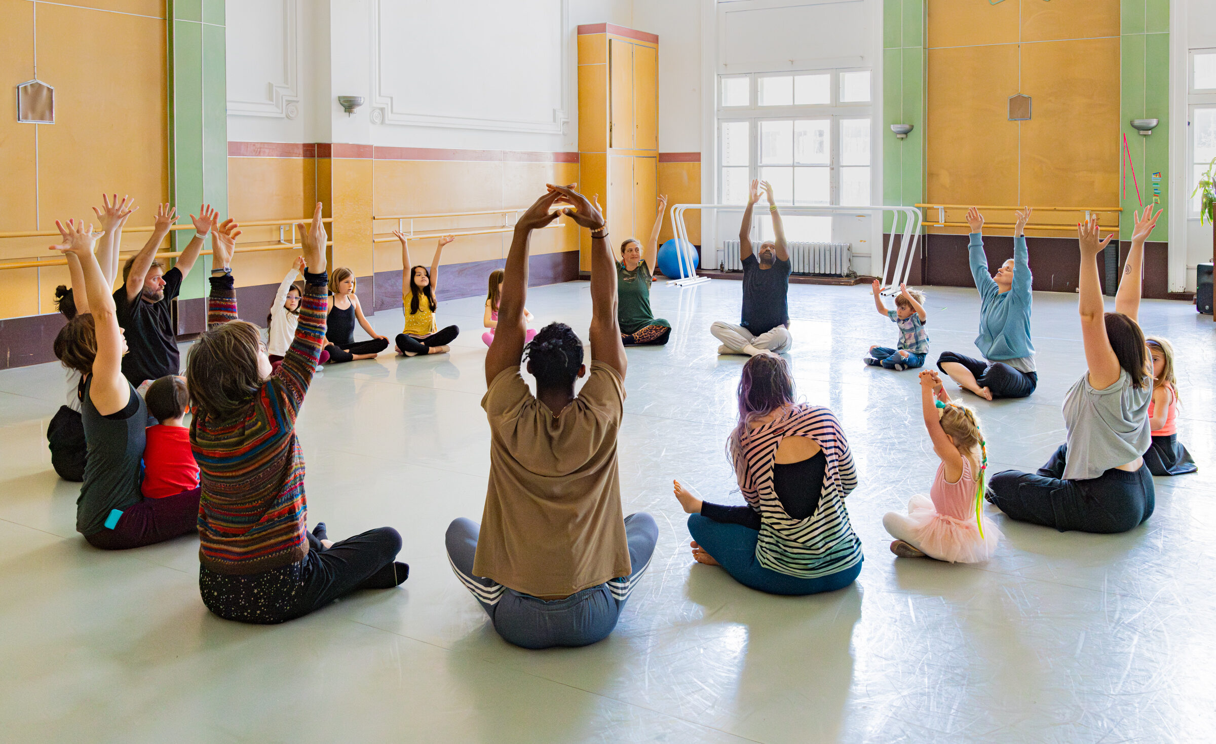 Family Workshop 2024 participants with LINES Community Programs Director Mary Carbonara and LINES Ballet company dancers Babatunji and Josh Francique, sitting in a circle stretching their arms upwards in a studio at LINES Dance Center