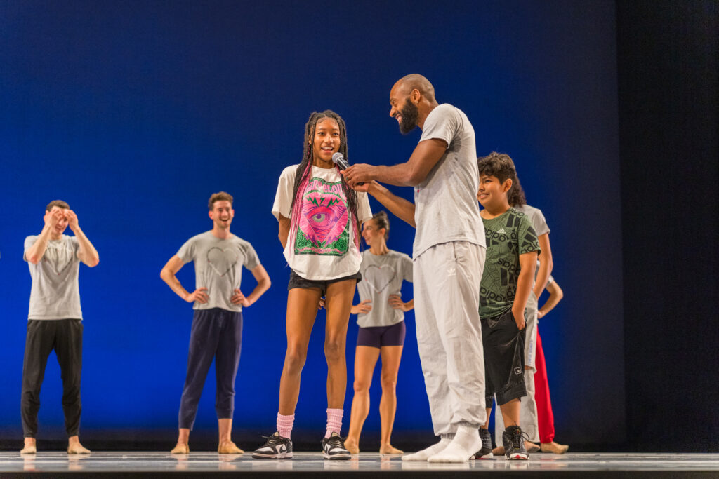LINES Ballet company dancer Babatunji holding a microphone and talking on stage with a student at a student matinee performance; company dancers are standing behind them on stage smiling