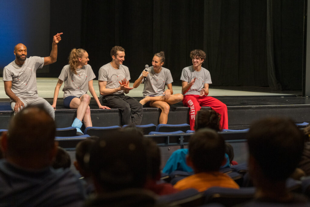 LINES Ballet company dancers sitting on stage during a performance for students; Ilaria Guerra is holding a microphone during a Q&A with the students