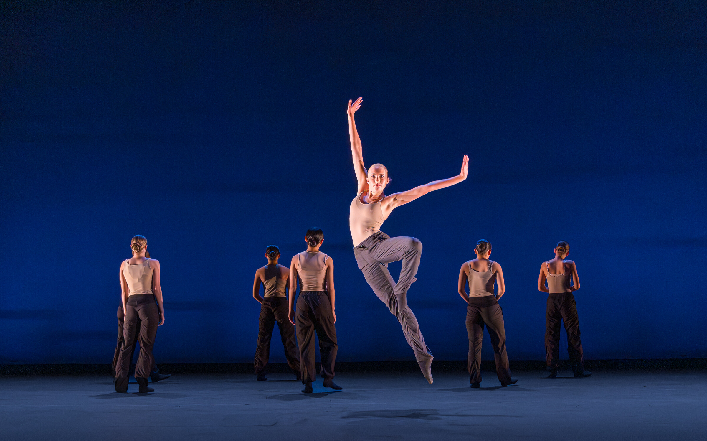 A dancer performing a piece by Carmen Rozestraten on stage in a Pre-Professional LINES Ballet Summer Program Showcase. Dancer in virtuosic jump wearing a skin toned top and black pants in front of a line of dancers standing with their backs to the audience.