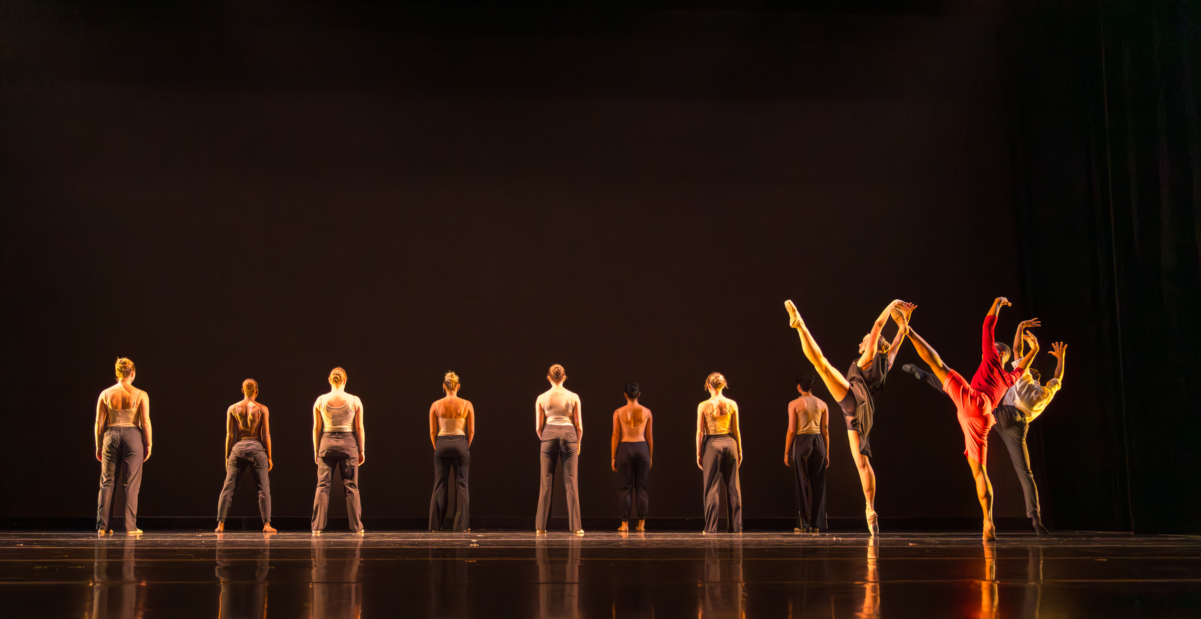 Three Summer Program dancers developpé their legs high to the side in front of a line of dancers facing the back of the stage