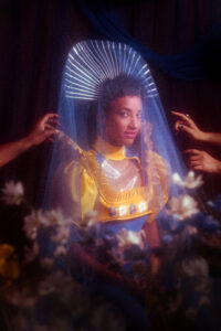 esperanza spalding looking at the camera in a gold and blue outfit with a sheer blue veil over an arcing headpiece