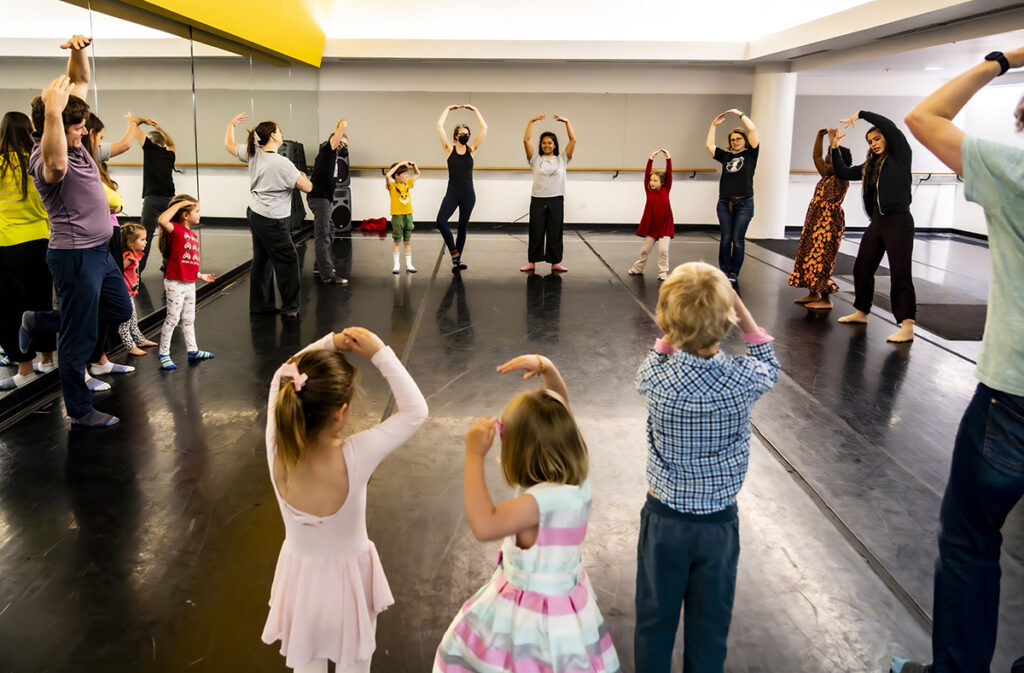 Kids and caregivers stand in a circle with their arms raised above their head, following a LINES Teaching Artist through a dance exploration during LINES Family Experience