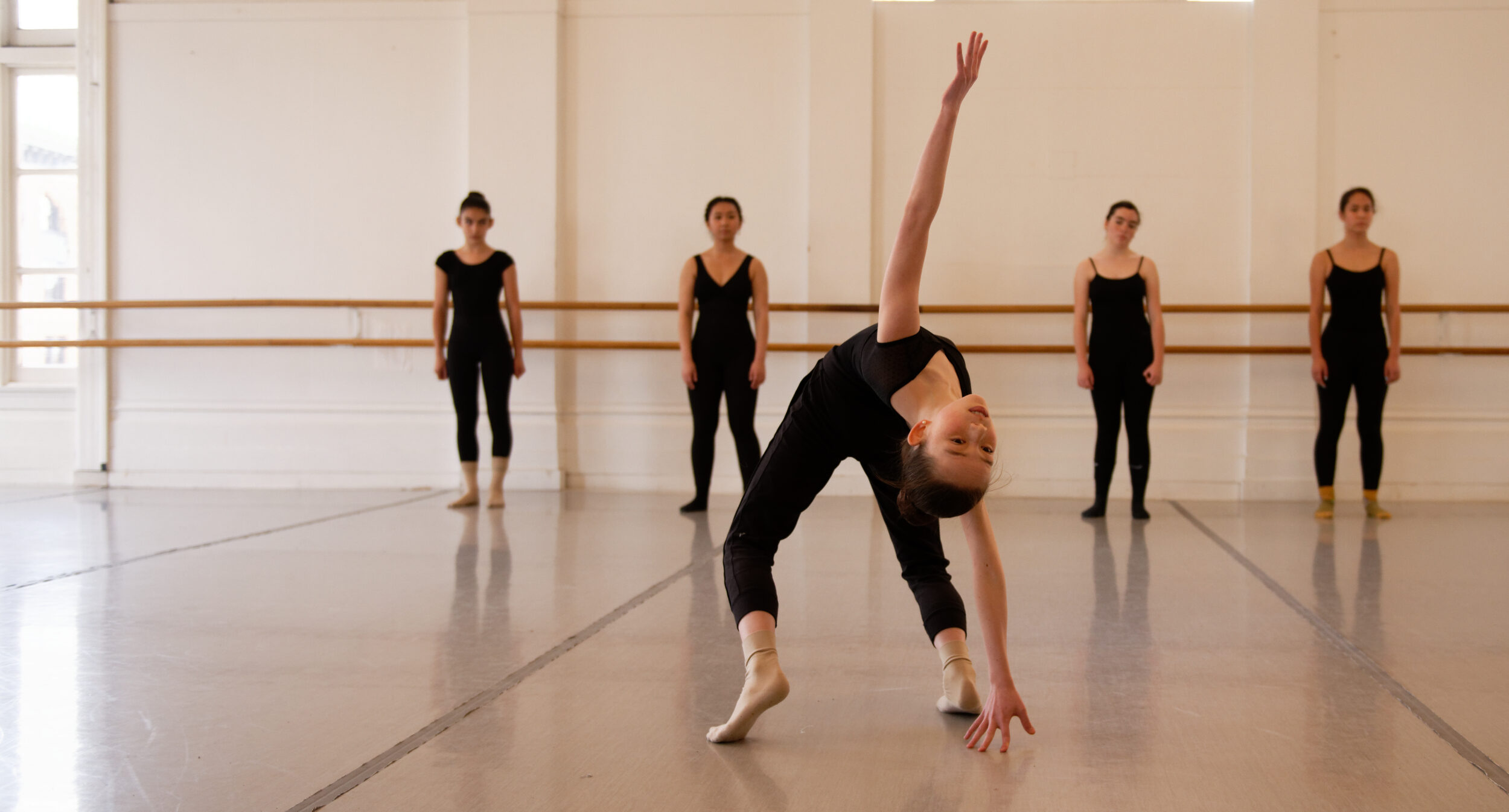 A young dancer in black dance clothes does a back bend while four others stand watching behind them.
