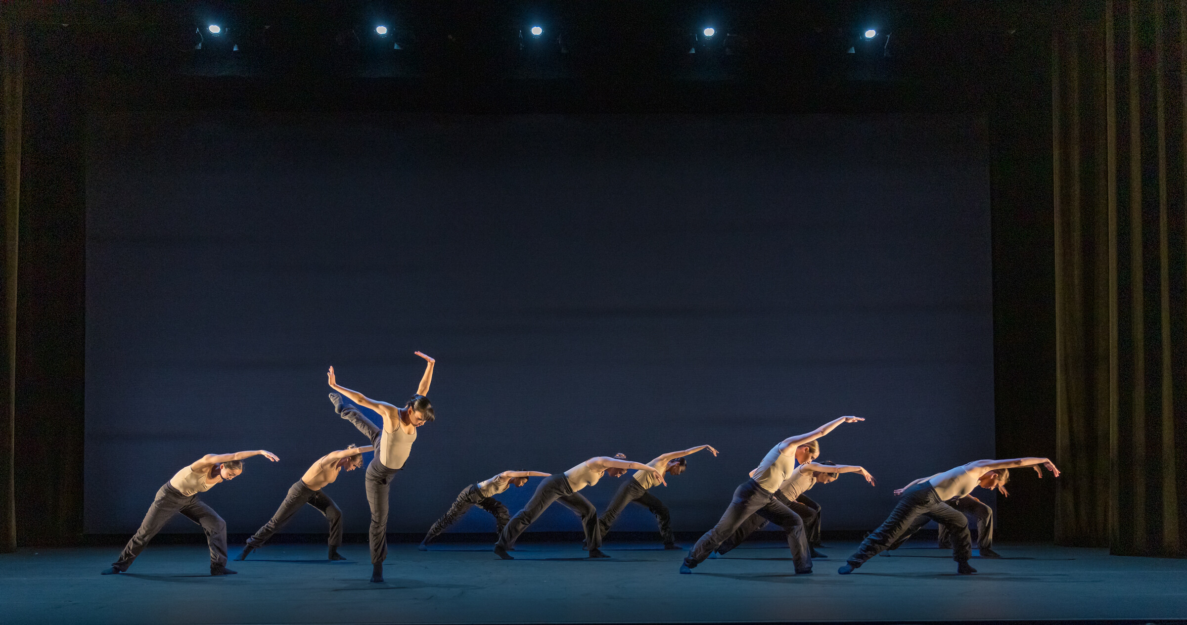Summer Program students perform onstage wearing black pants and skin toned tops, a group of them bent to the side in unison with one dancer in an impressive back attitude in the front