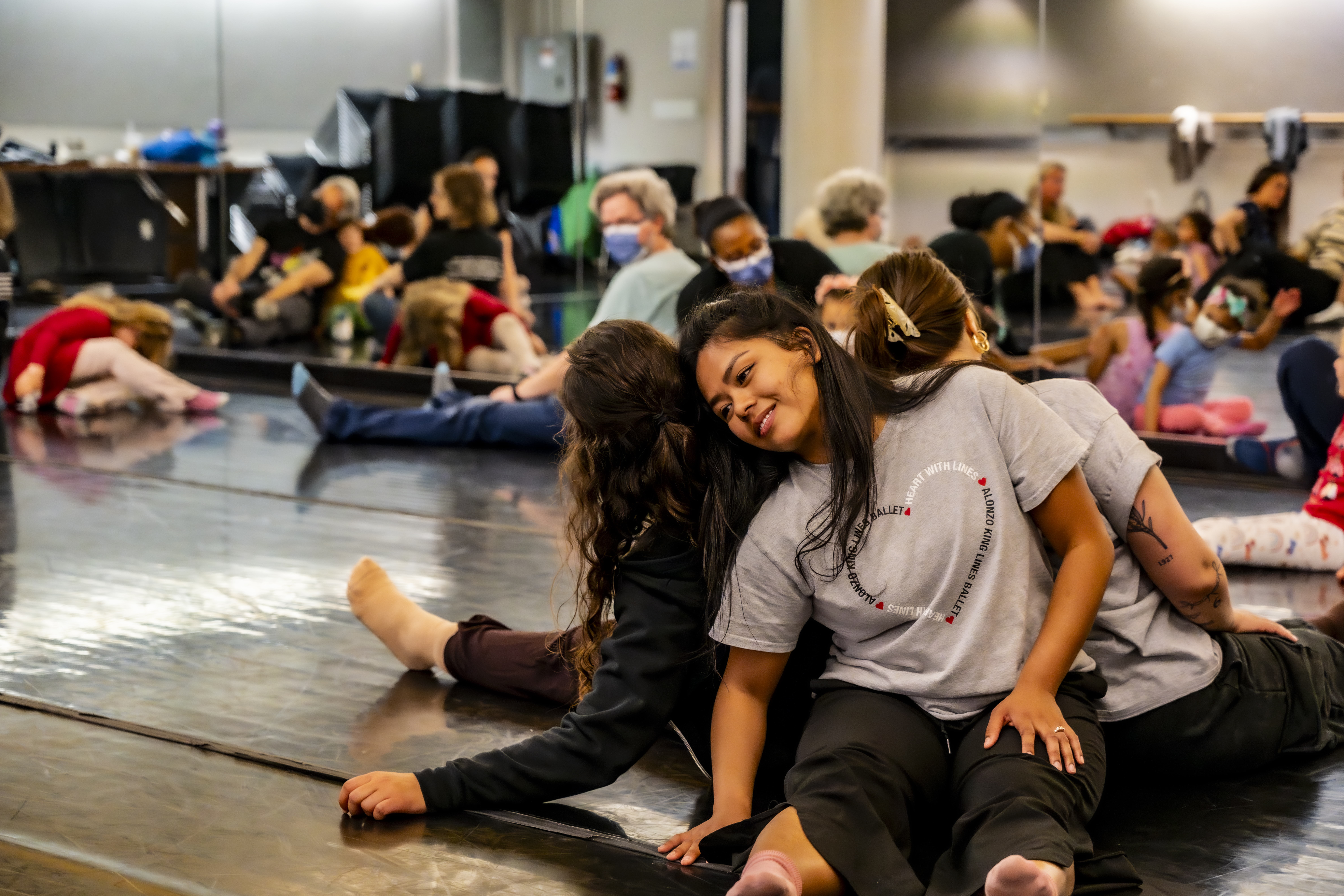 Kids and caregivers sit back to back in a circle, following a LINES Teaching Artist through a dance exploration during LINES Family Experience