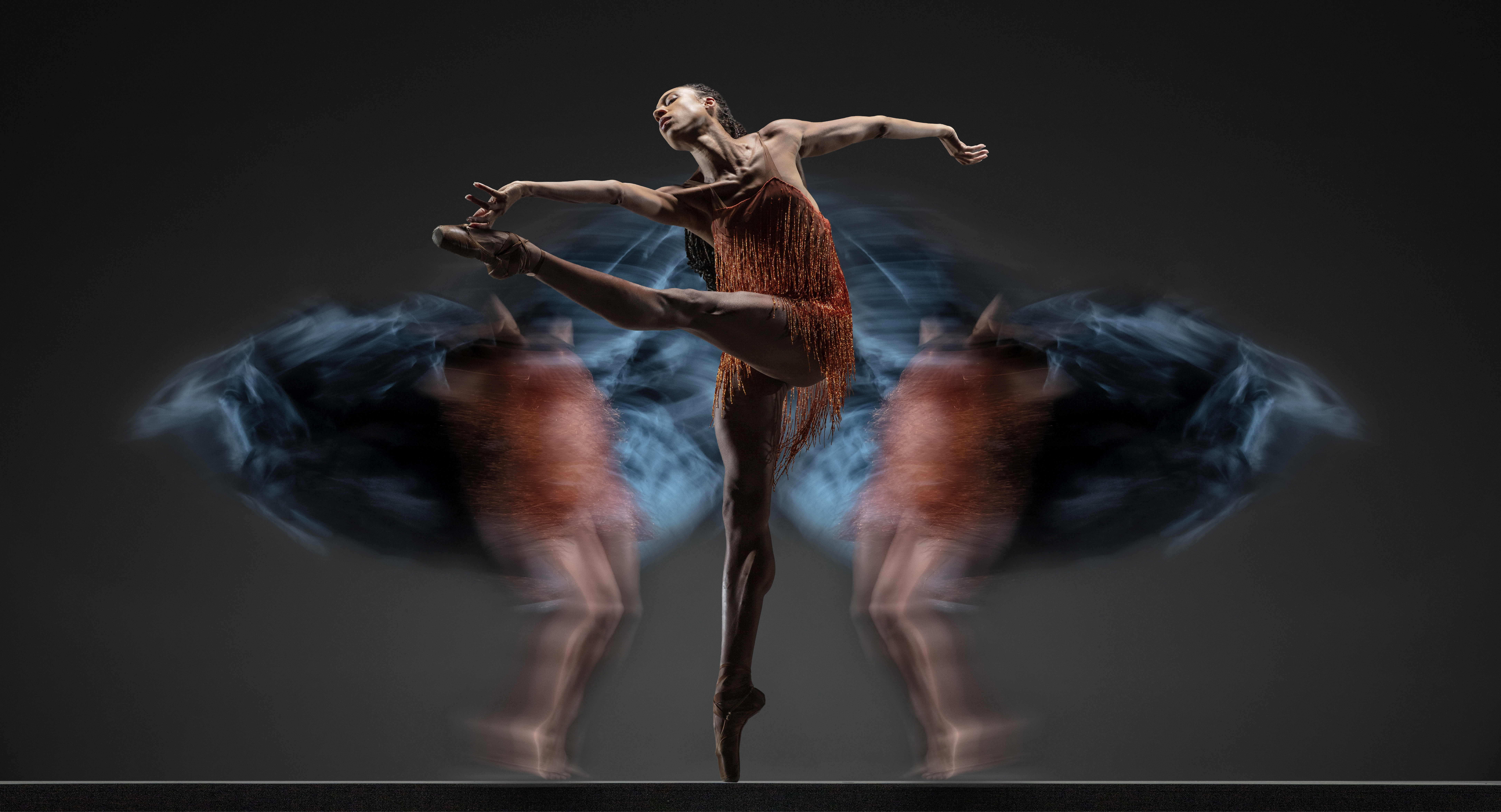 Company dancer Adji Cissoko reaches towards one foot lifted to the front wearing a beaded orange, sparkly costume and surrounded on either side by blurred dancers also in orange and teal.
