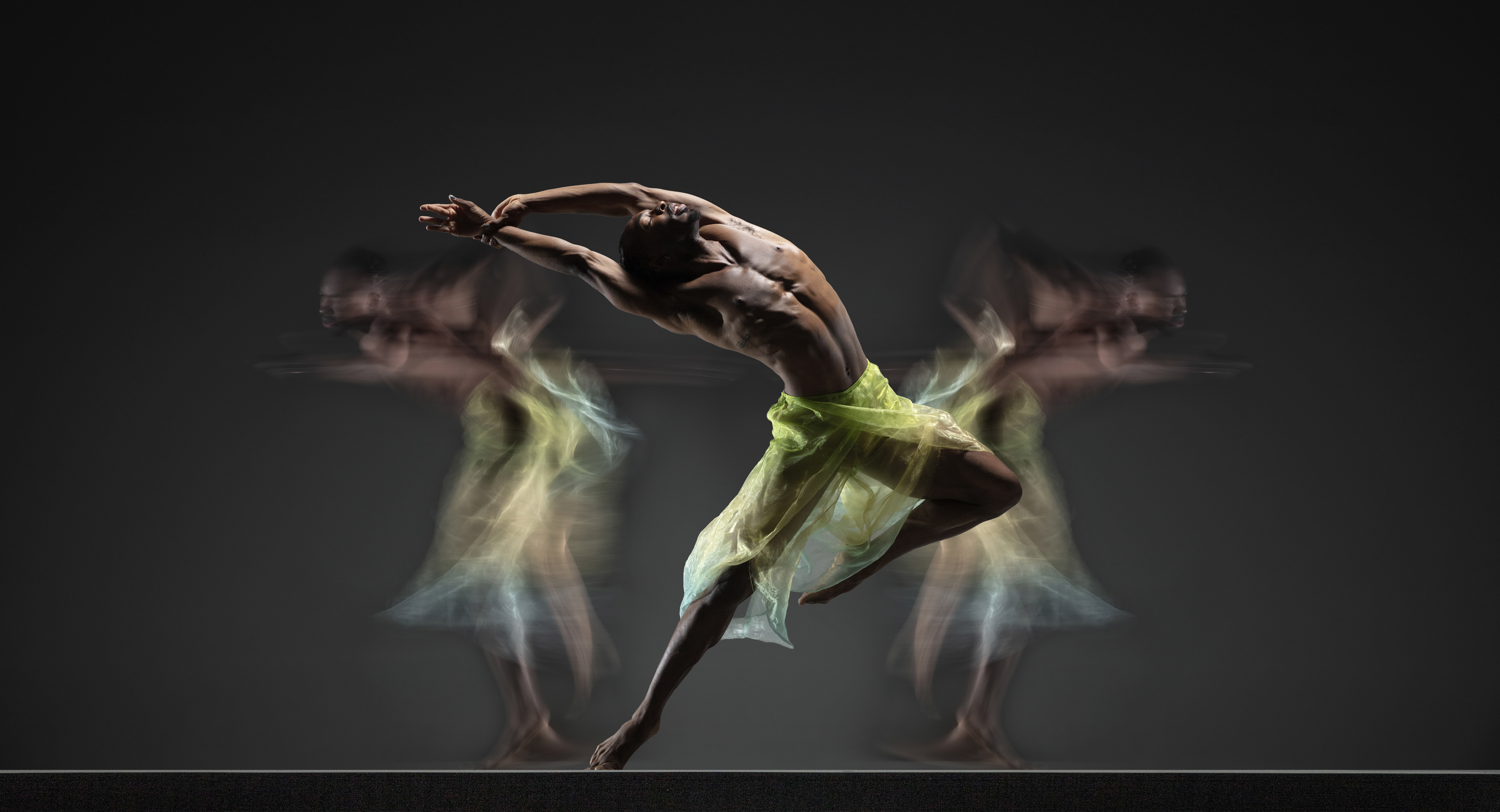 Company dancer Josh Francique arches back in a daring balance on one foot, face to the sky, wearing flowy green and teal pants. Two blurred bodies in the same pants move in the background.