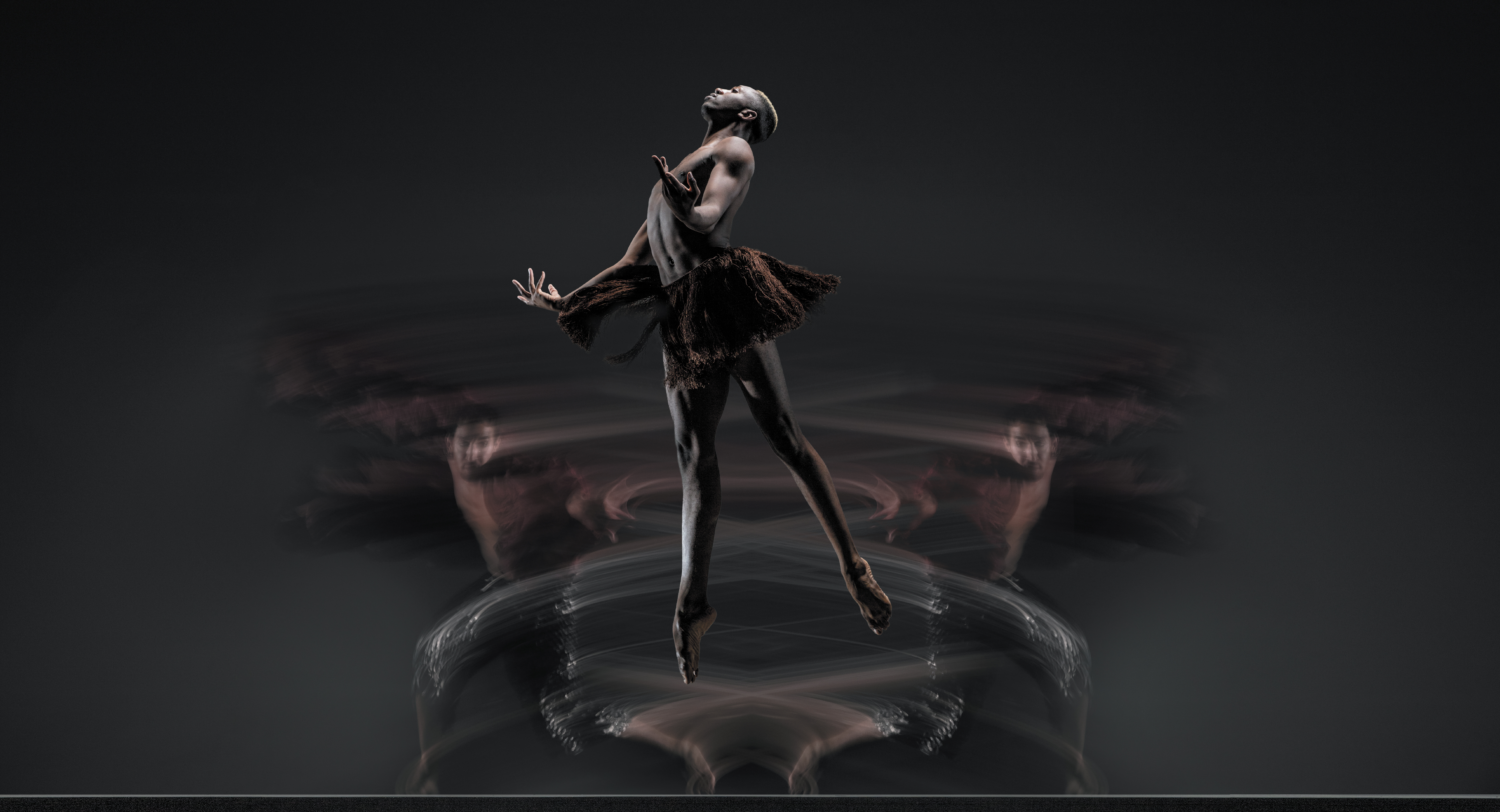Company dancer Mikal Gilbert jumps with arms open and legs slightly split, wearing a short brown skirt. Two blurred bodies move in the background.