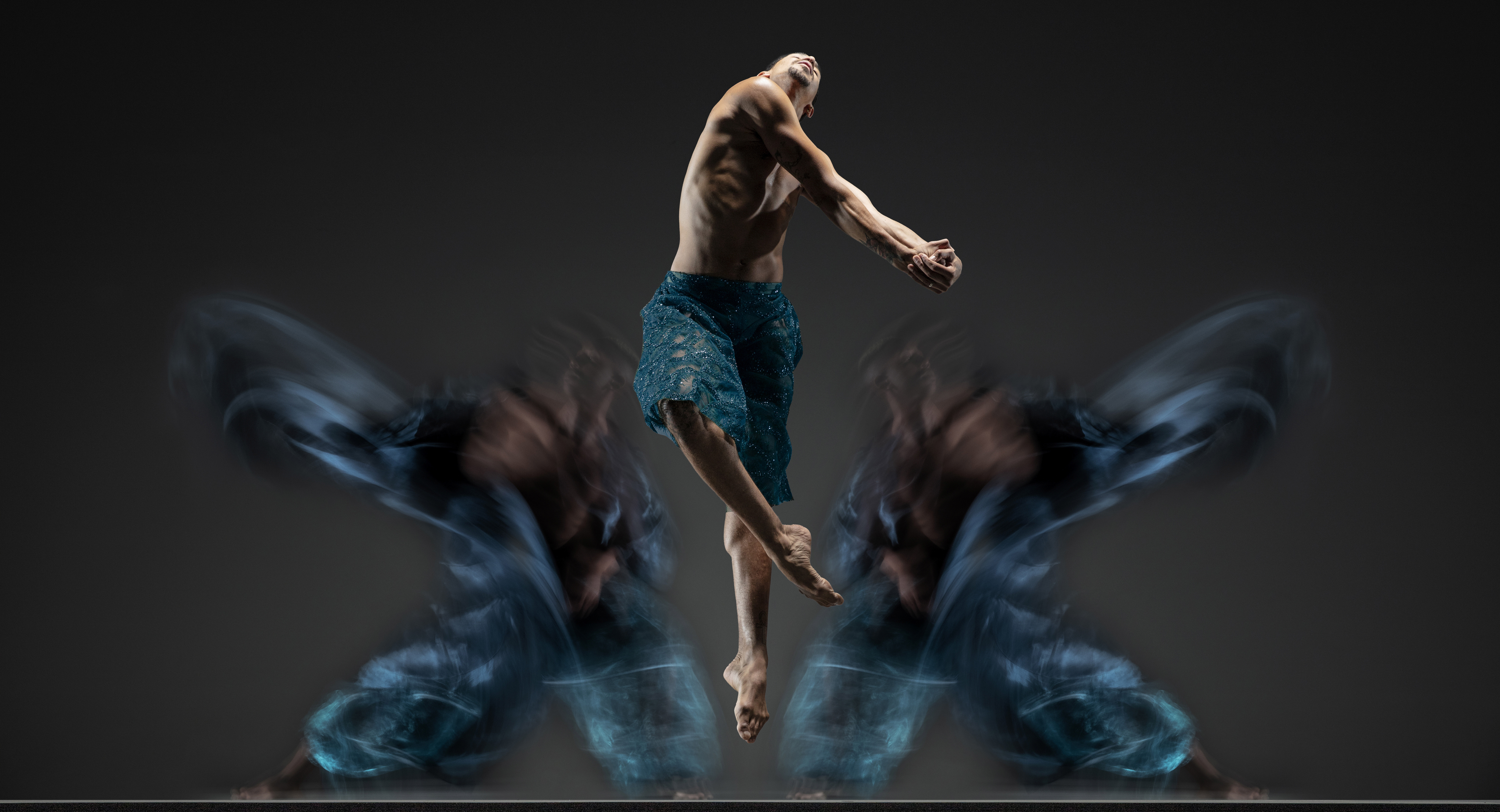 Company dancer Shuaib Elhassan jumps with arms and legs spiraling, wearing intricate teal shorts. Two blurred bodies in teal move in the background.