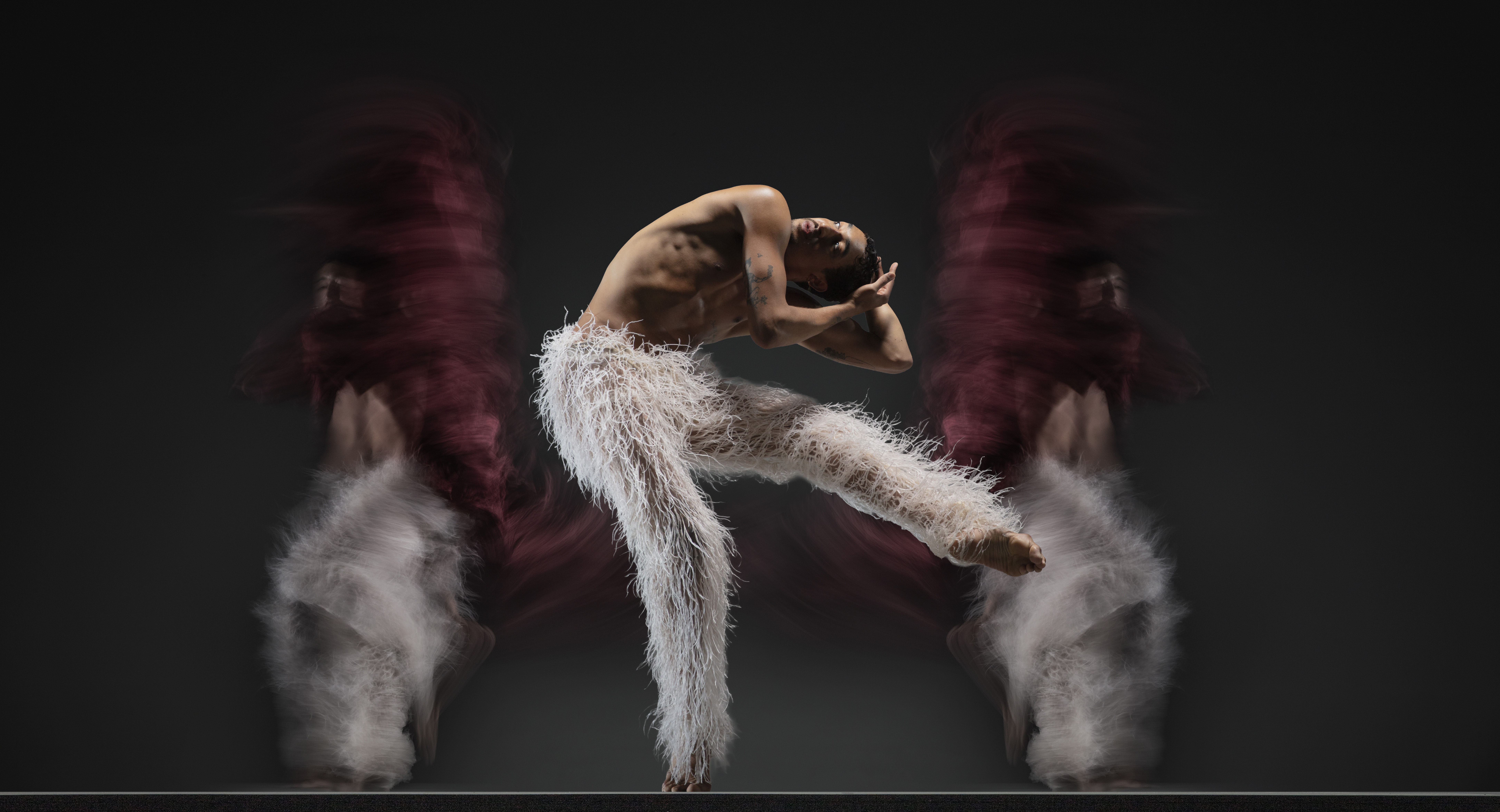 Company dancer Shuaib Elhassan folds over holding his head with one leg up, wearing white pants with long white strings. Two blurred bodies in white and burgundy move in the background.