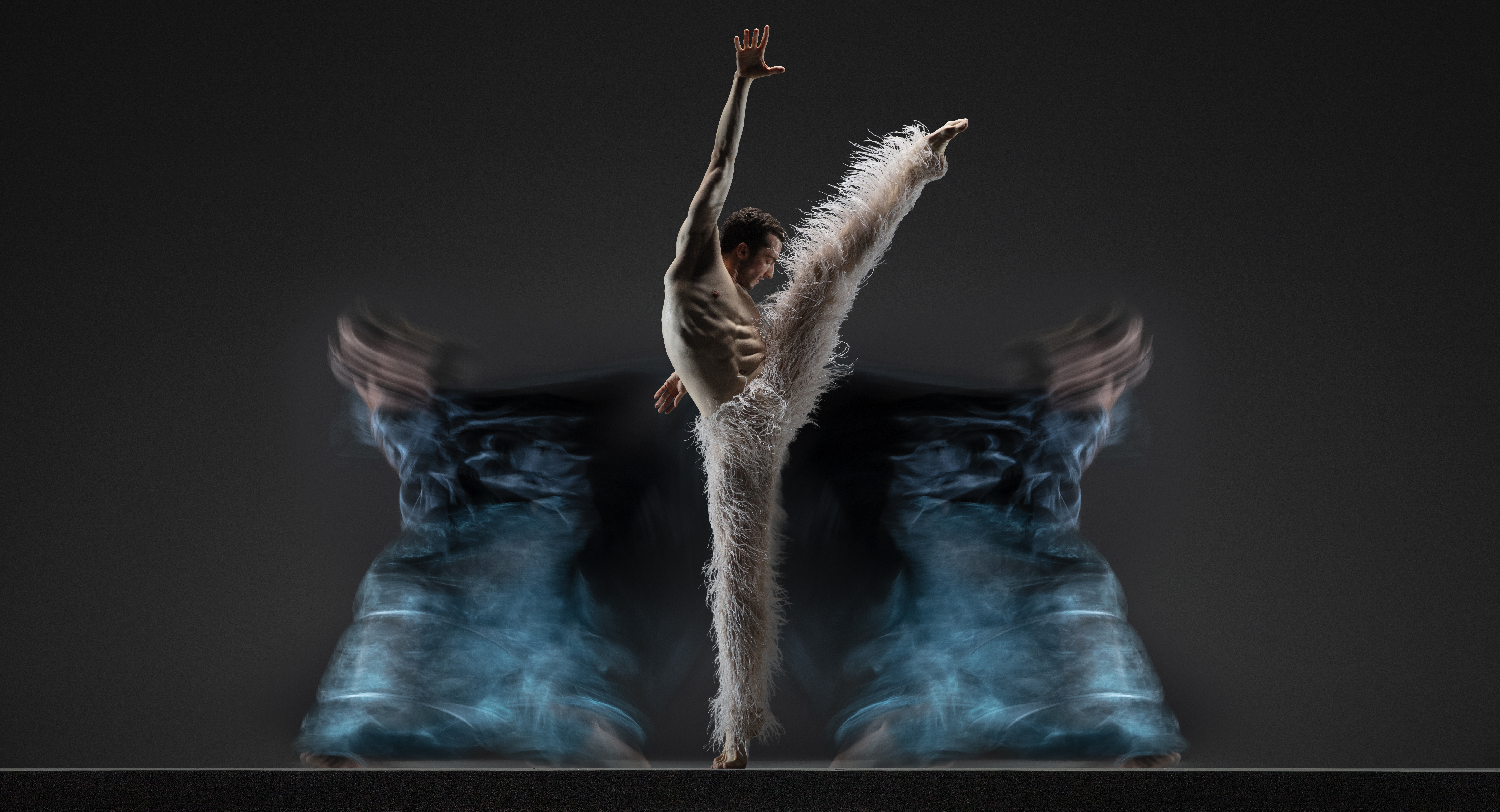 Company dancer Theo Duff-Grant lifts his leg in a high developpe front reaching his opposite arm up in opposition, wearing white pants with long white strings. Two blurred bodies in teal move in the background.