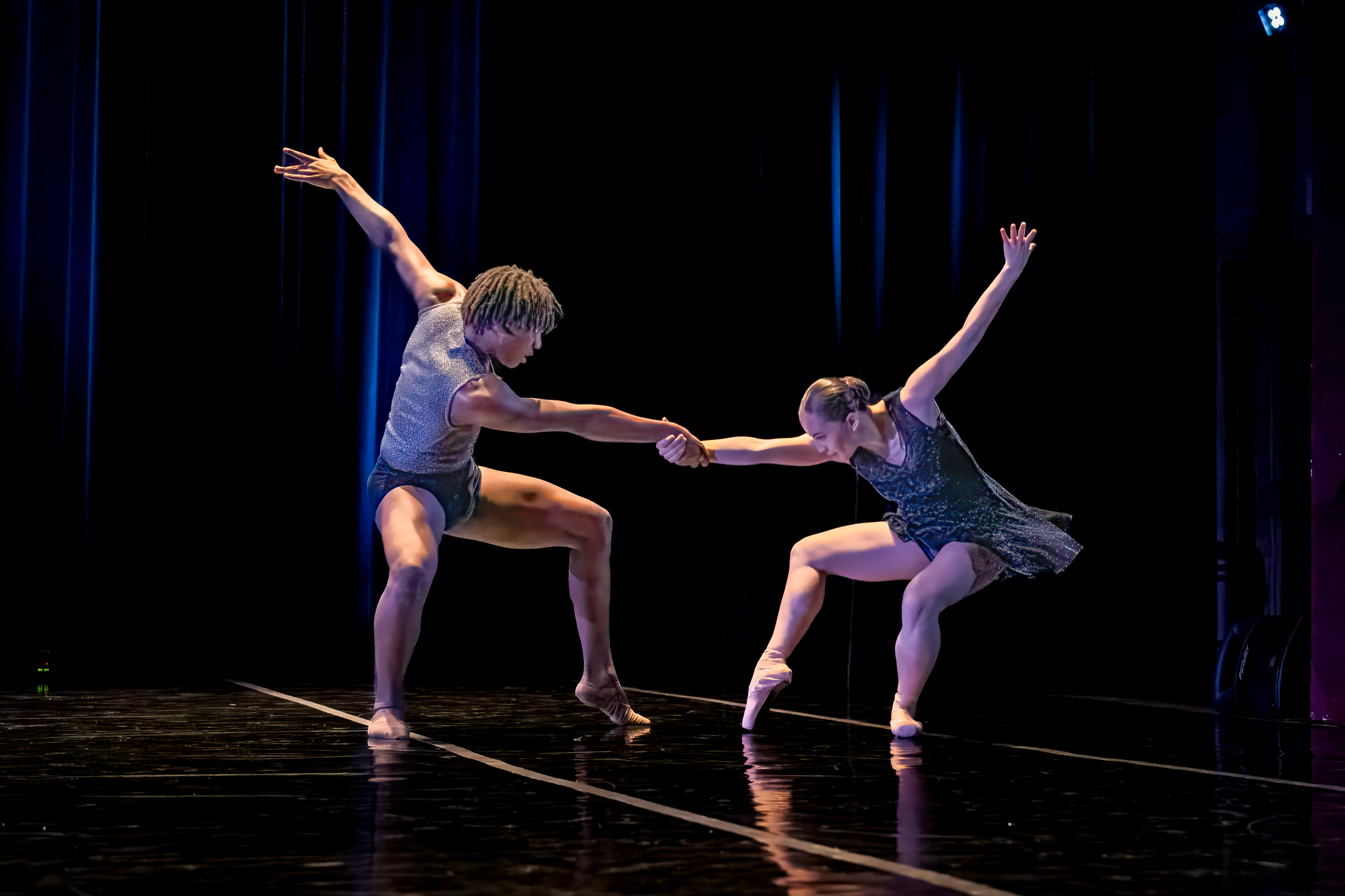Two dancers in gray costumes pull against each other in counterbalance, one foot extended in front of each with a pointed bevel.