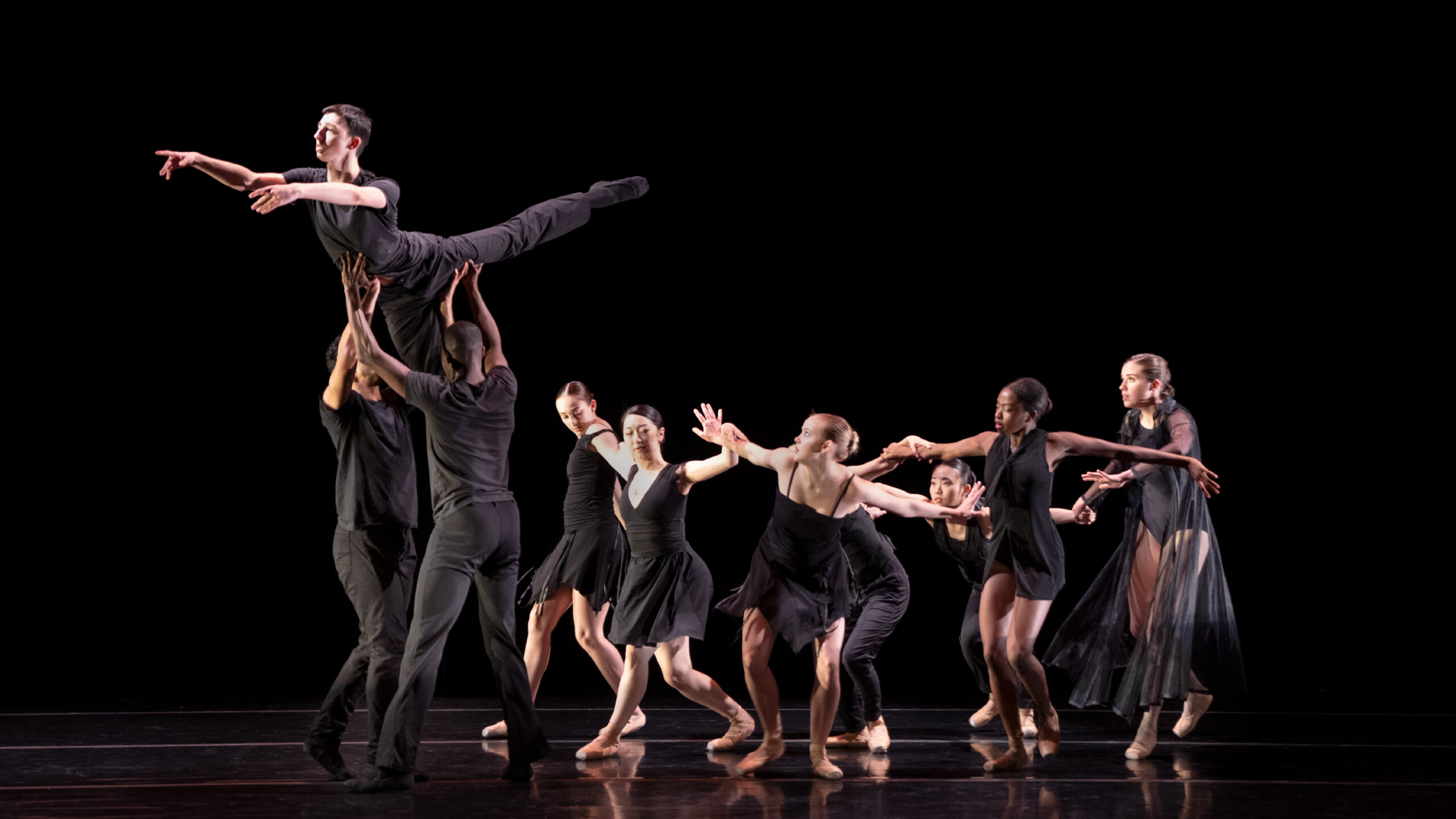 Summer Program dancers perform onstage in black costumes, one lifted and reaching by two others, as a group follows and watches.