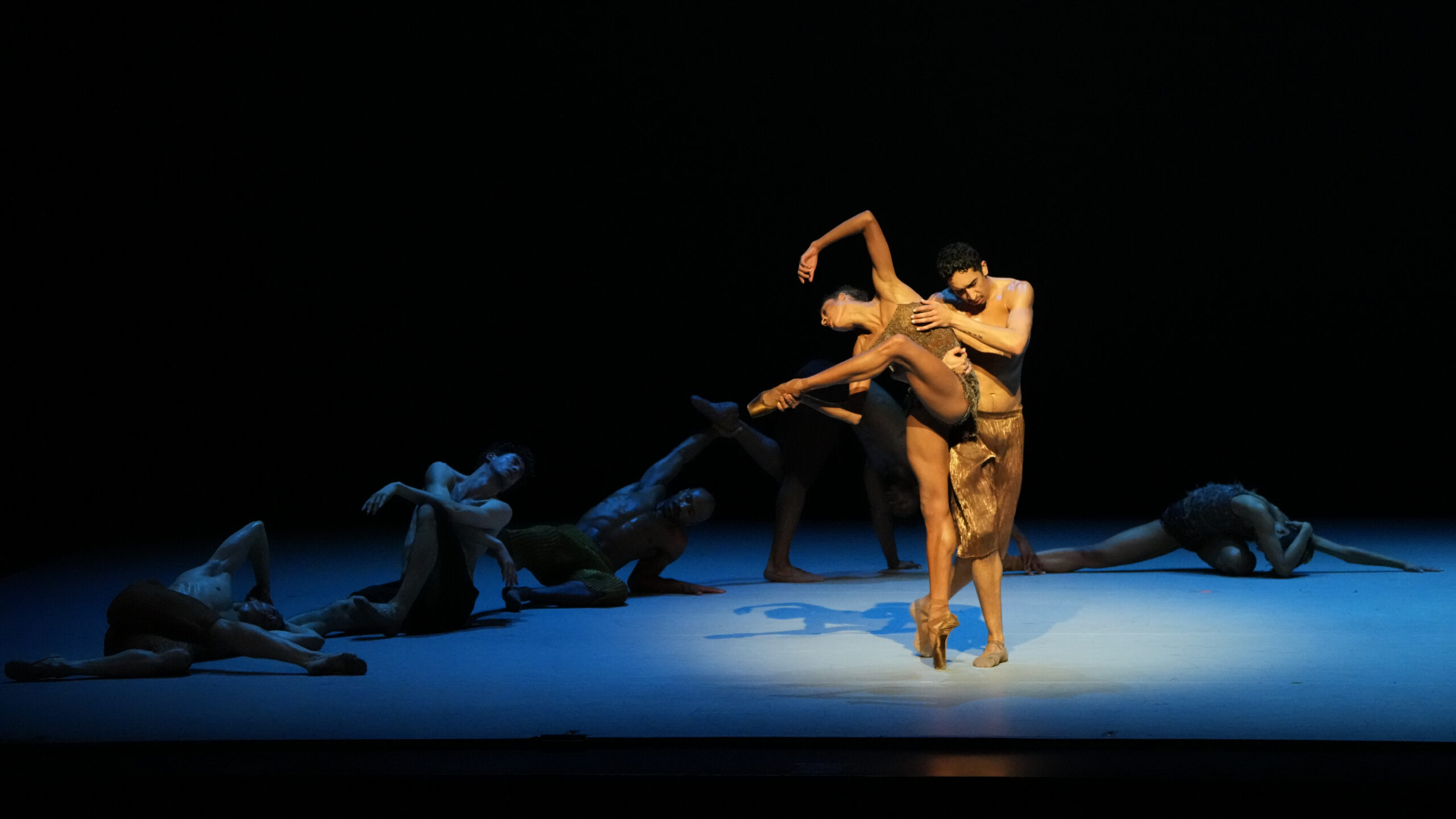Company Dancers Adji Cissoko and Shuaib Elhassan perform in 'Deep River' along with the LINES Company. Adji is balanced on one leg leaning over, supported by Shuaib, both wearing gold costumes.