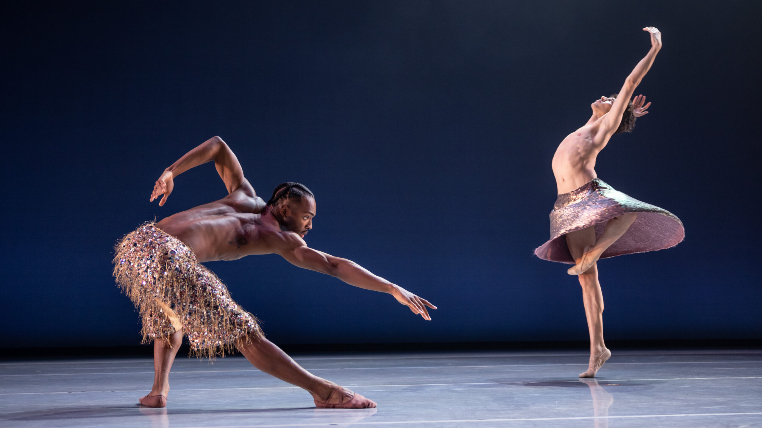 Company Dancers Joshua Francique and Maël Amatoul perform together onstage. Joshua wears textured pants and is in a deep lunge reaching towards the floor while Maël wears a metallic skirt that swings as he turns and reaches towards the sky.