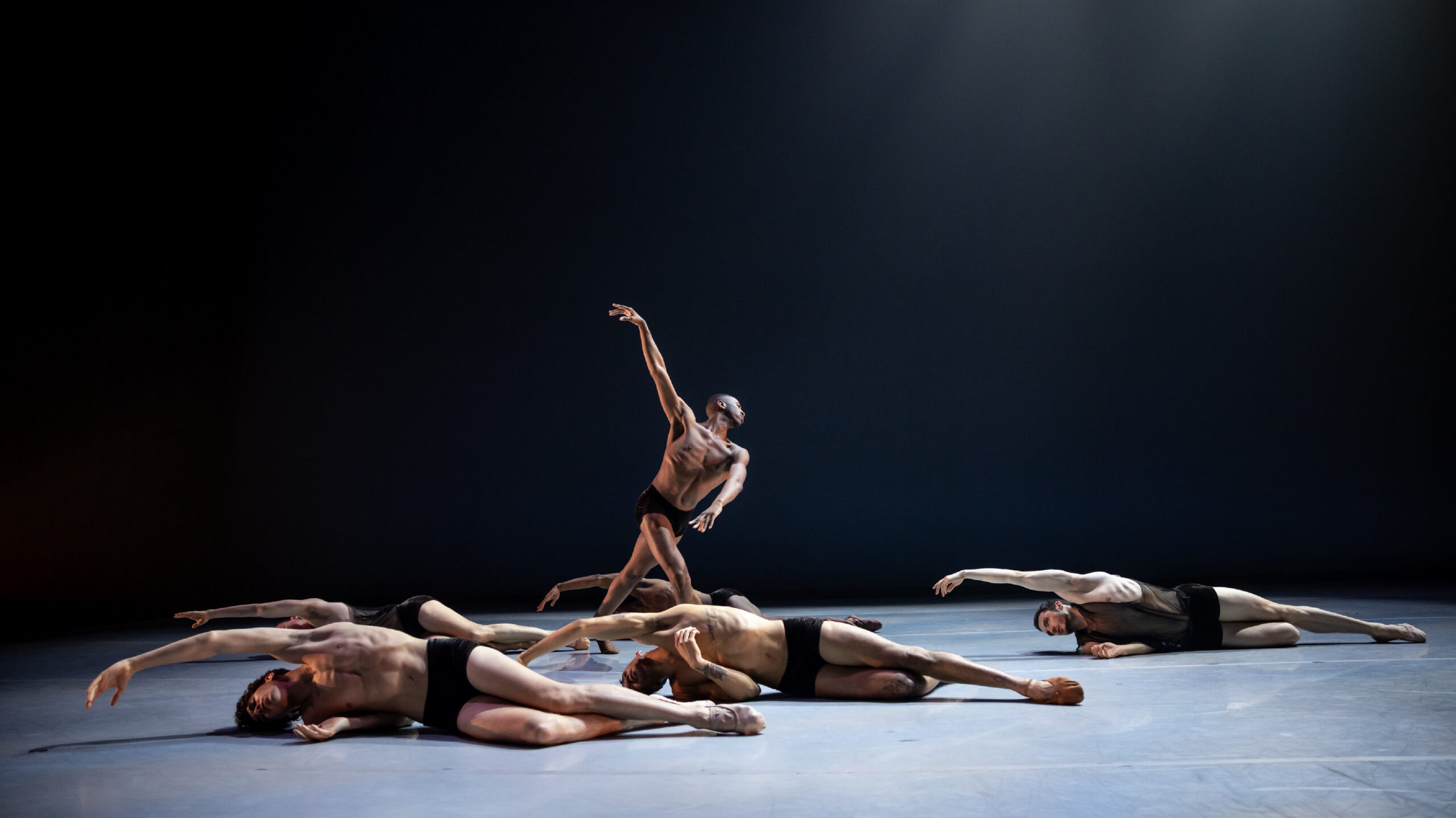 Company Dancer Mikal Gilbert bends to the side, standing over company dancers reaching to on the floor.