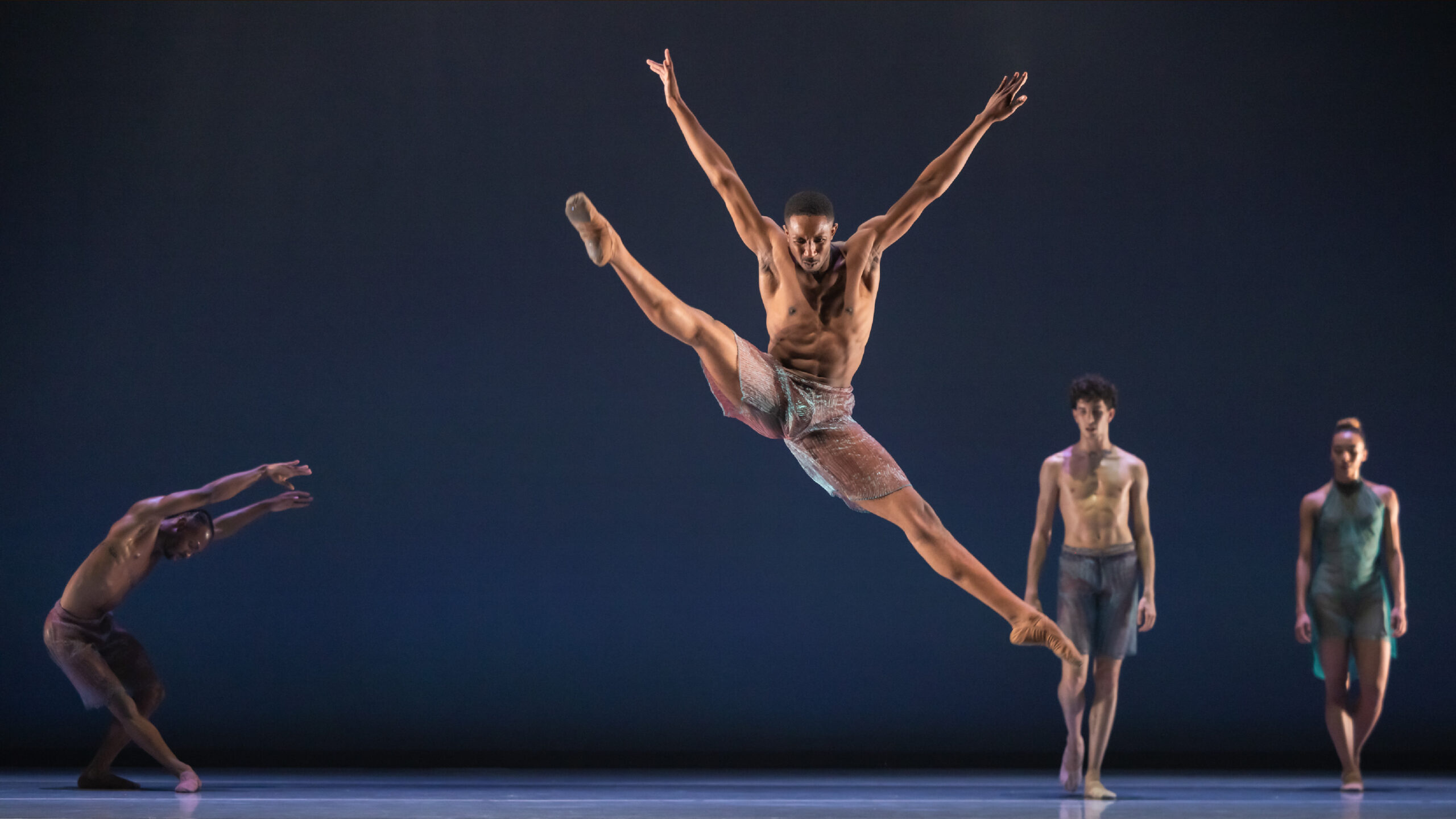 Company Dancer Mikal Gilbert jumps in a full split wearing colorful, shiny pants, sharing the stage with the LINES Company.
