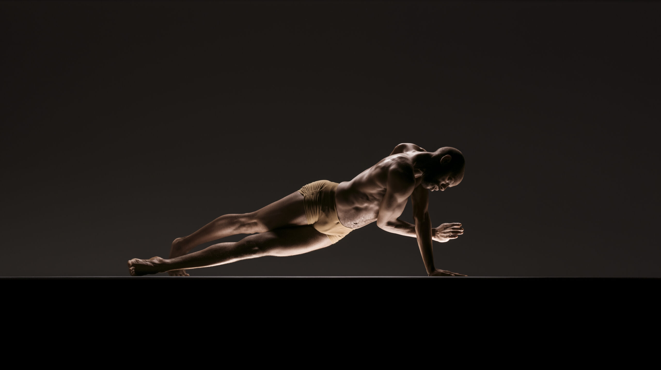 Former LINES Ballet Dancer Keelan Whitmore balances in a plank on one hand, focus towards a light source on the floor, otherwise surrounded by darkness.