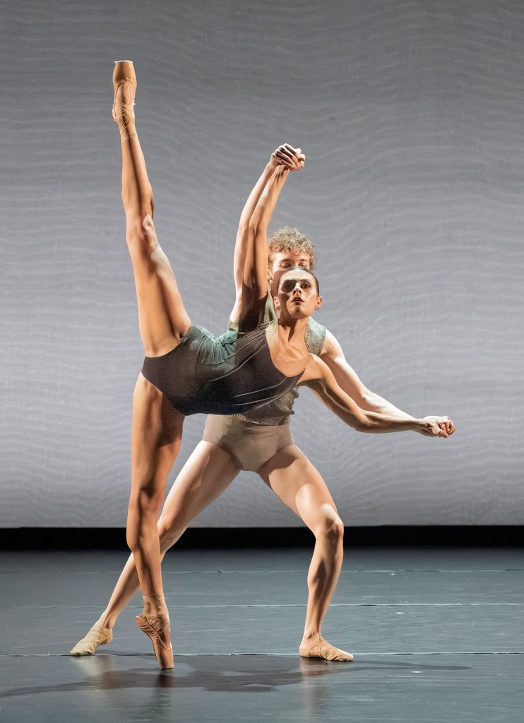 2 LINES Summer Program dancers perform onstage in gray leotards/unitards. One dancer supports the other en pointe in a split penche.