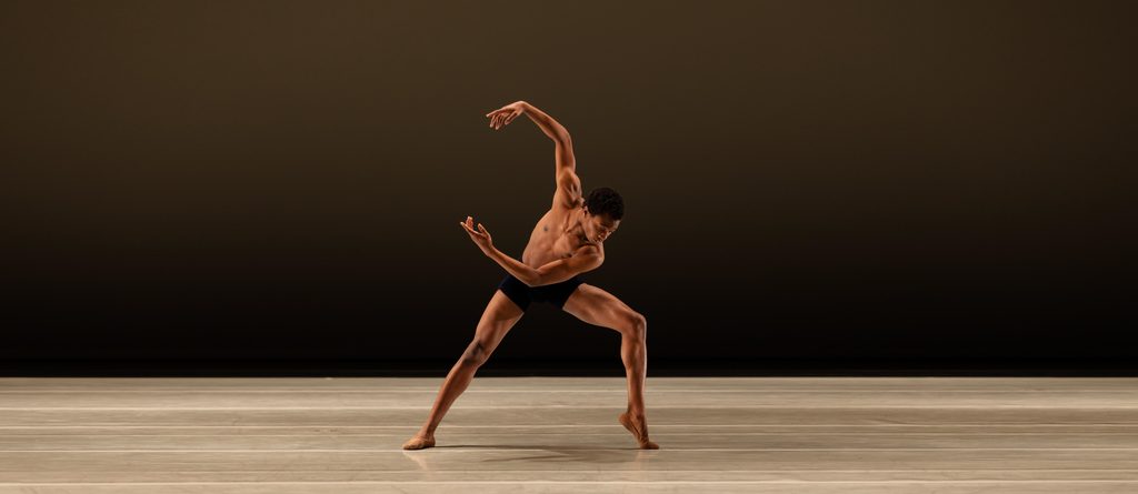 LINES Education dancer moves onstage, wearing black bike shorts, arms rounded to the side and in a forced arch on one foot.