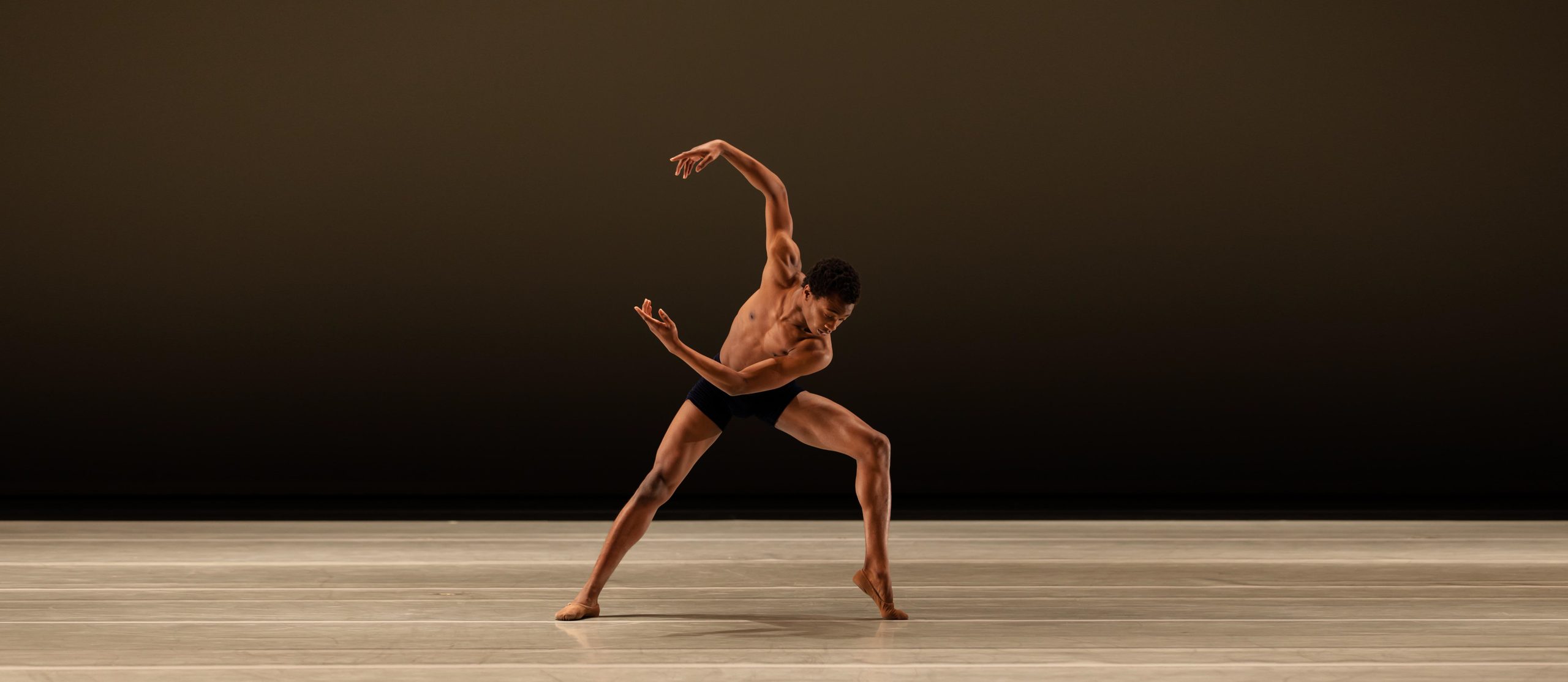 LINES Education dancer moves onstage, wearing black bike shorts, arms rounded to the side and in a forced arch on one foot.