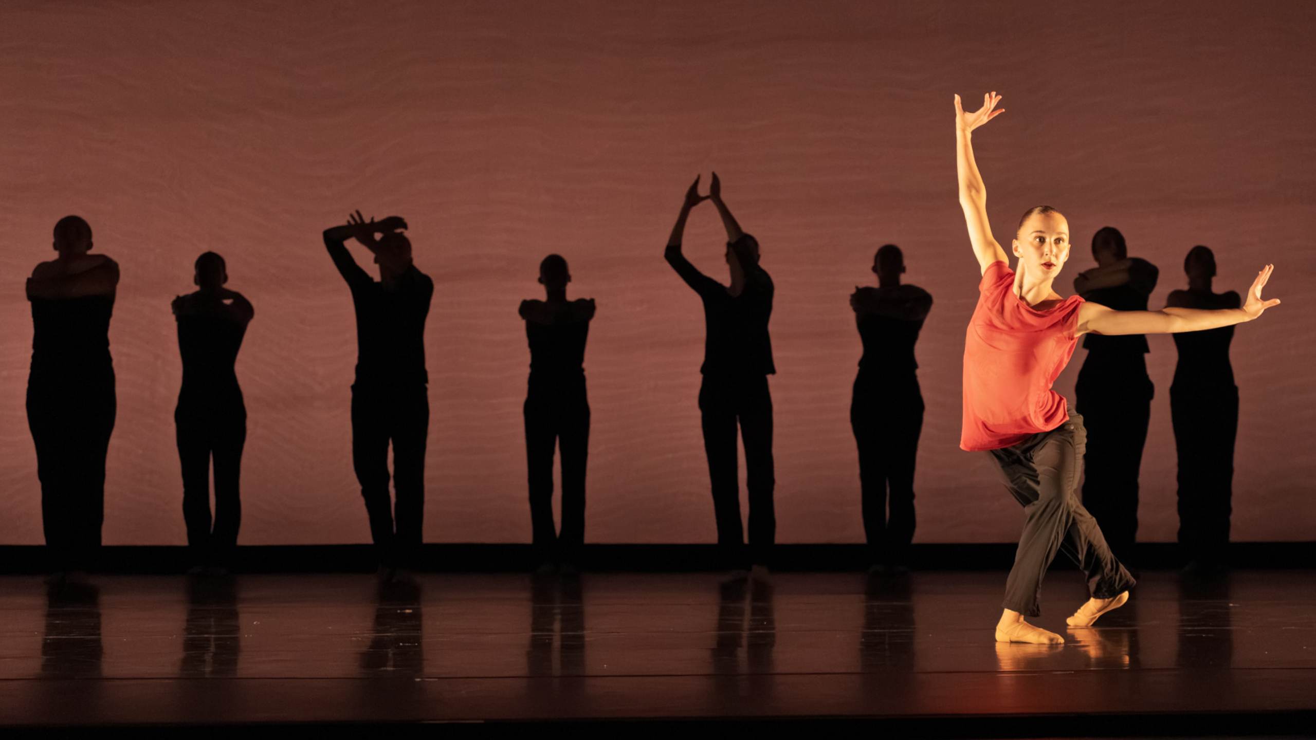Summer Program students perform onstage. A line of dark silhouettes against a deep brown backdrop stand behind a dancer in red top and black pants, braced in a 4th position with arms reaching up and to the side.