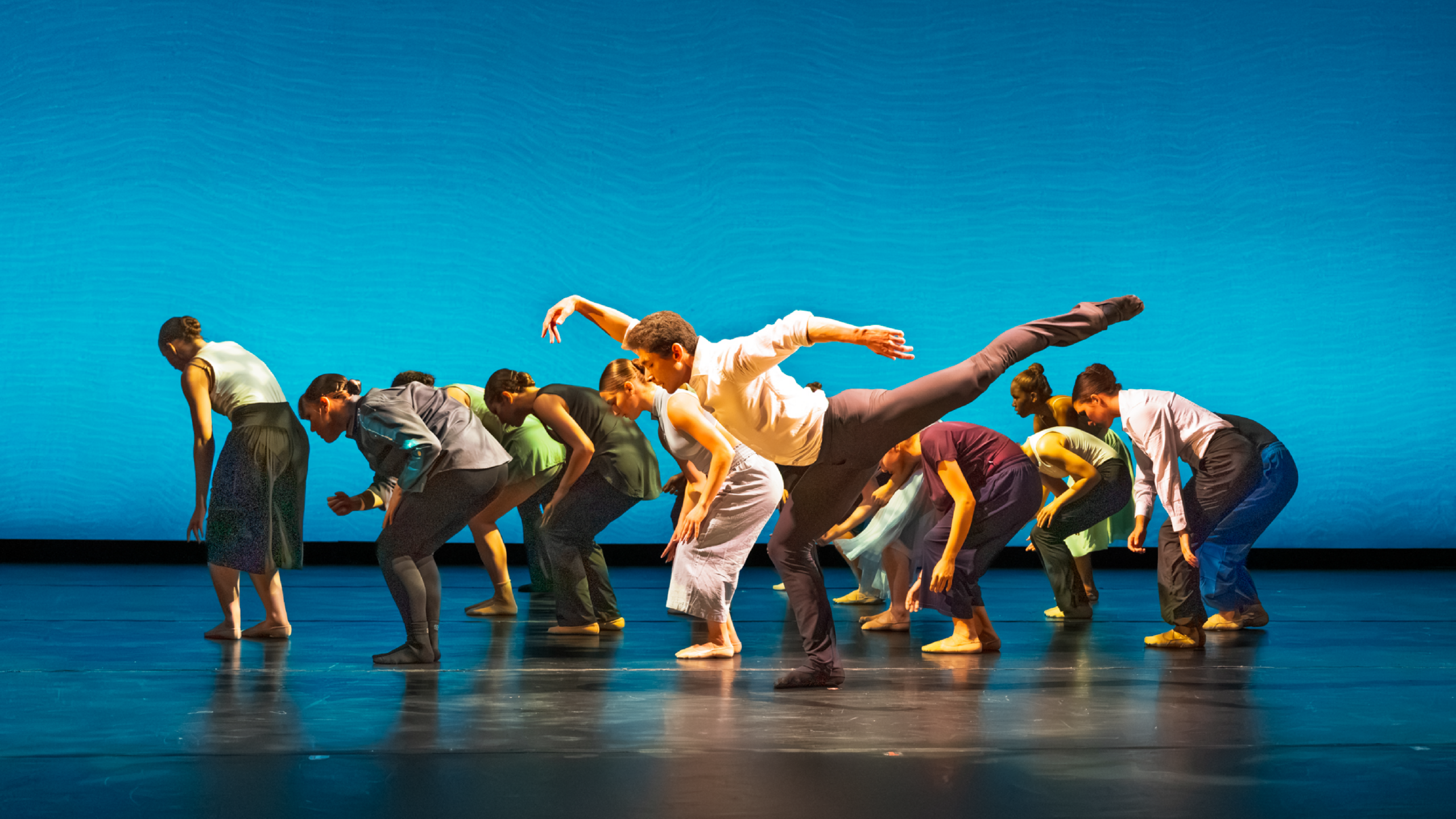 Summer Program students perform onstage with a teal backdrop. A group of dancers dressed in bright tops and black pants crouch down behind a dancer in a white button-down and black pants stretched in a long arabesque.
