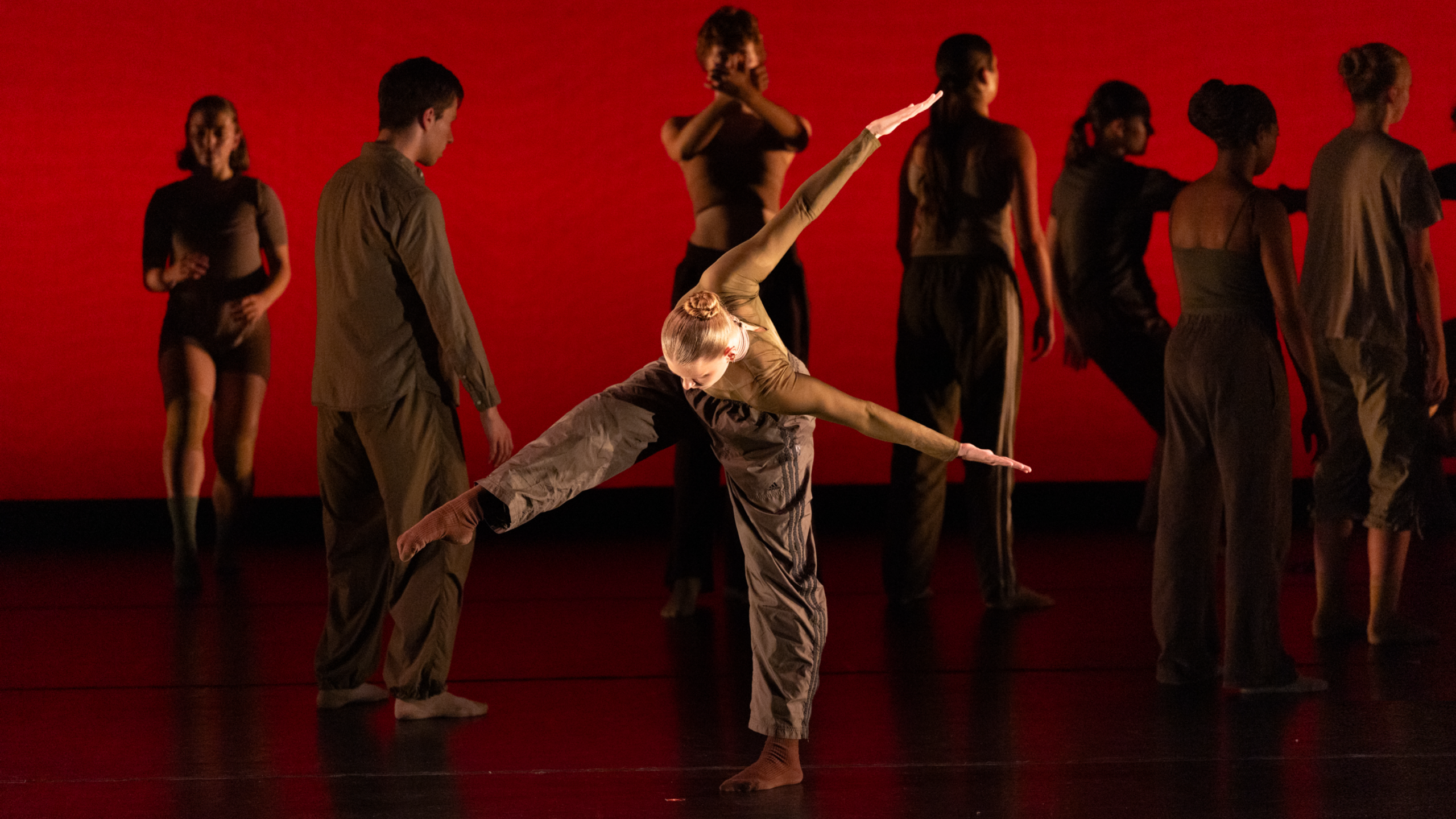 Summer Program students perform onstage. Dark silhouettes stand against a red backdrop, while one dancer dressed in browns at the forefront bends in half, arms behind their back, and one leg pointed out.