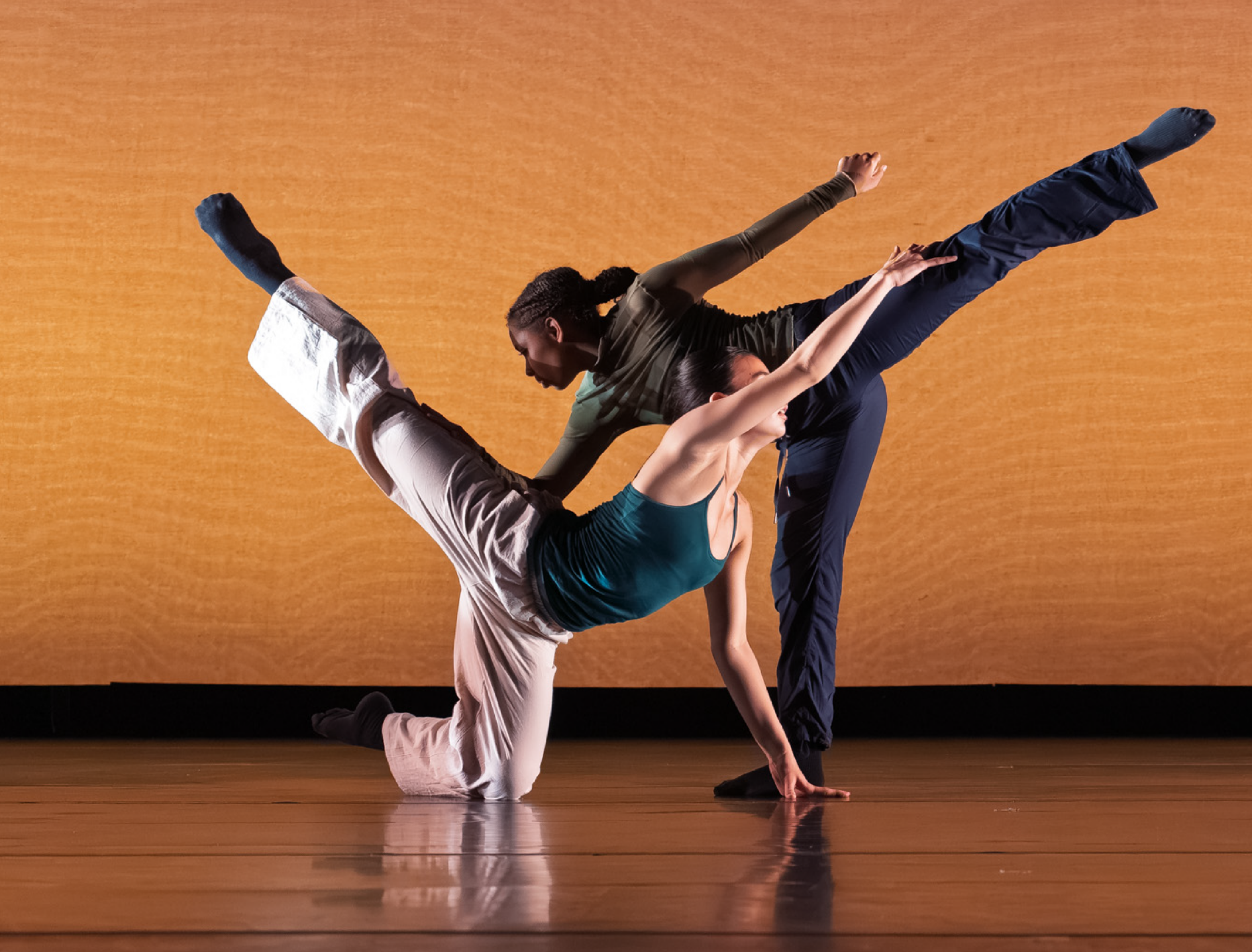 Summer Program students perform onstage. Two dancers on different levels support each other, both with legs reaching high behind them, as a third watches in the background. Dancers are in neutral costumes against a light orange backdrop.